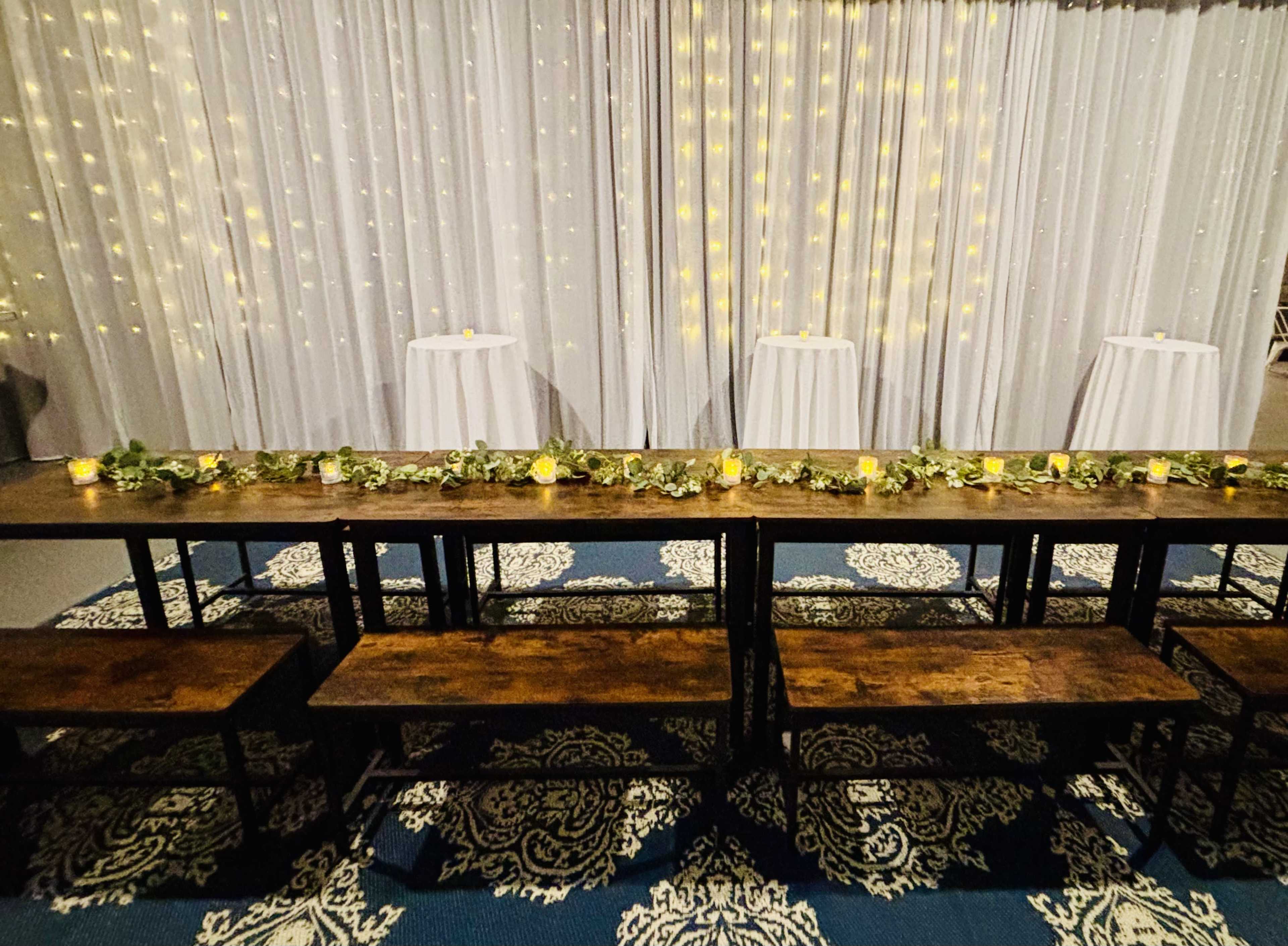 The image shows a decorated indoor venue with a long wooden table adorned with greenery and candles, surrounded by tall cocktail tables against a backdrop of sparkling fairy lights.