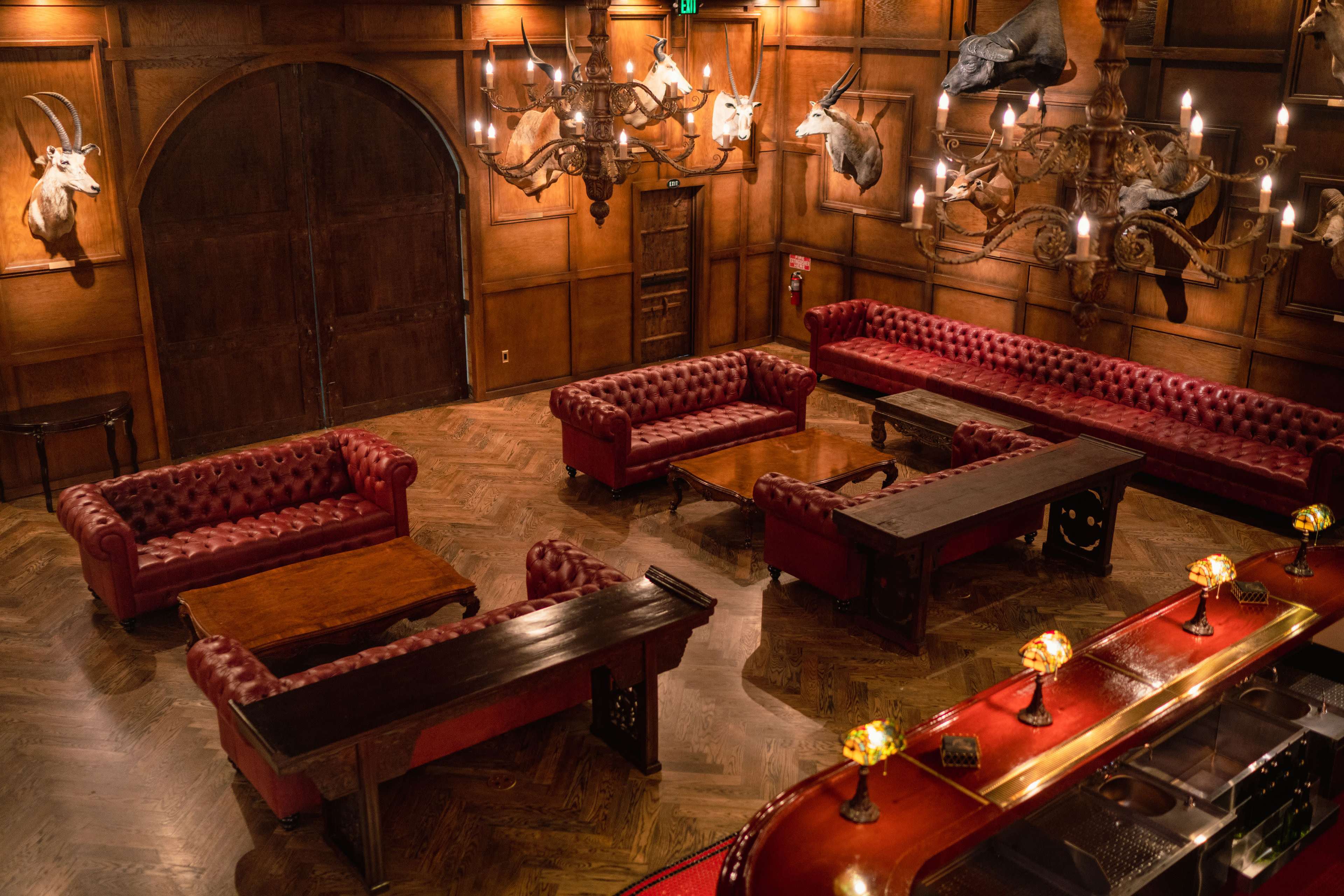 A dimly lit room with wooden walls, featuring red upholstered sofas, wooden tables, and mounted animal heads on the walls.