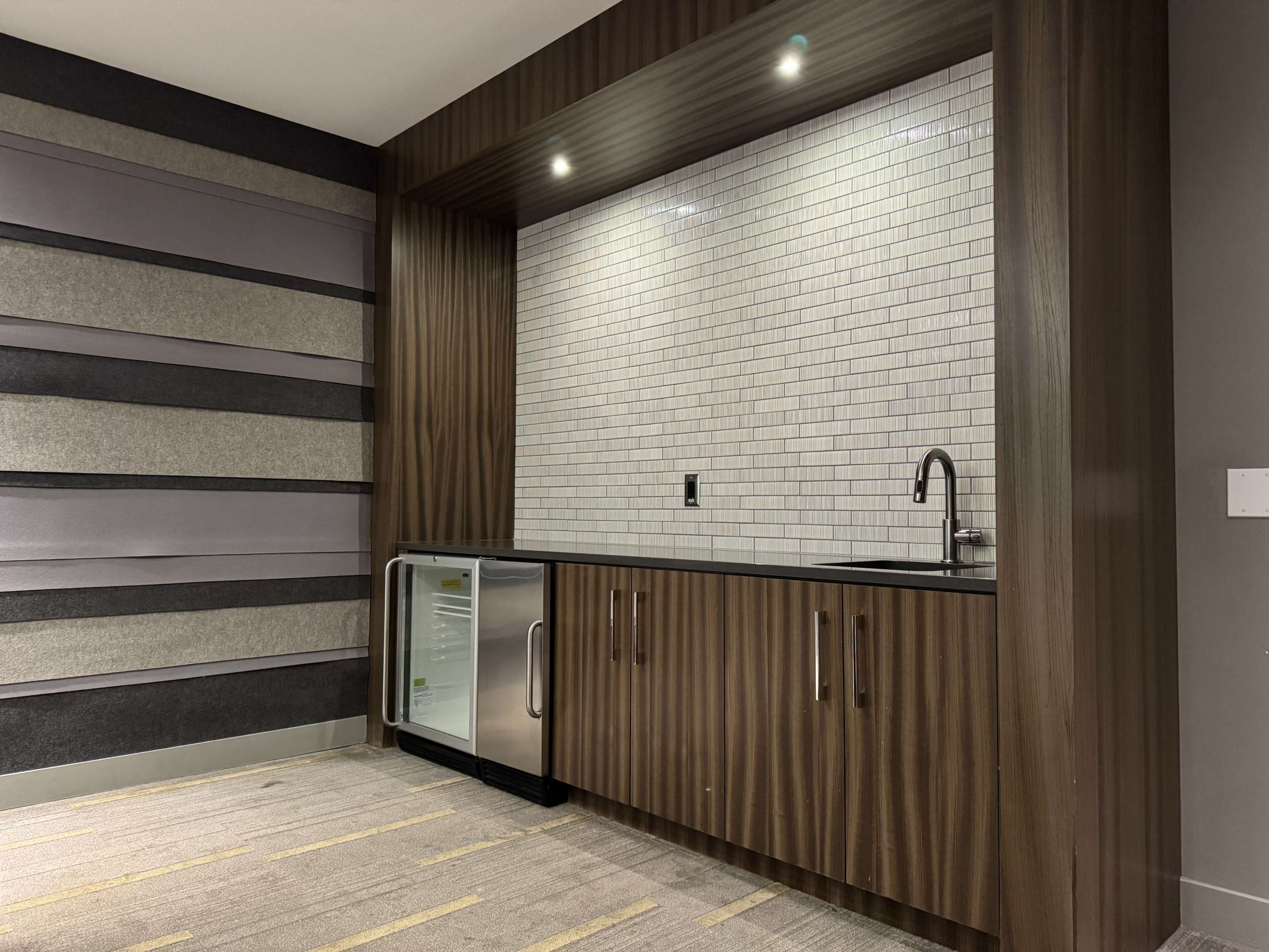The image shows a modern kitchenette with wood cabinetry, a sink, and a small refrigerator, set against a wall of textured panels in neutral tones.