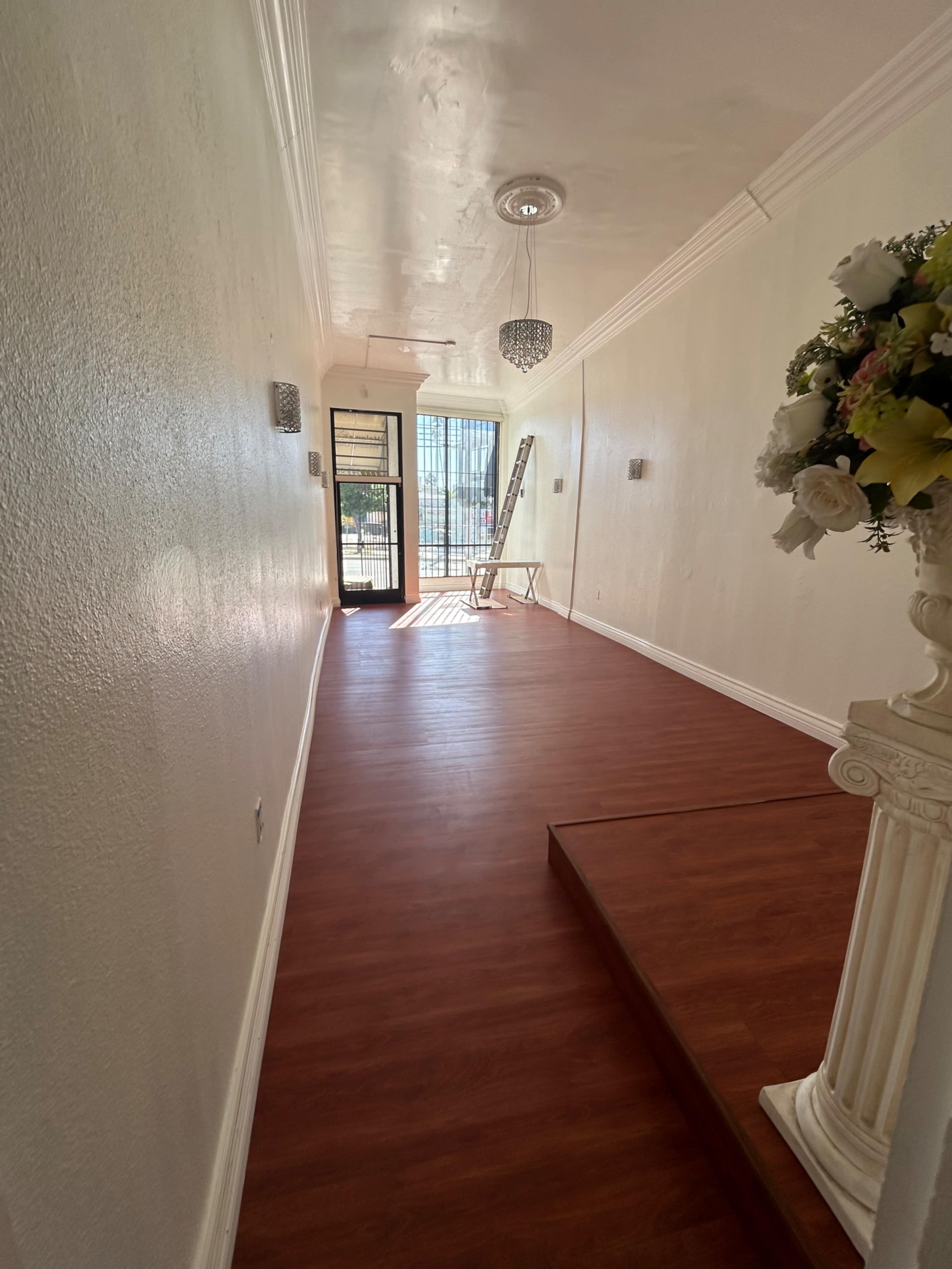 The image shows a well-lit hallway with wooden flooring, leading to a glass door at the end and adorned with decorative elements.