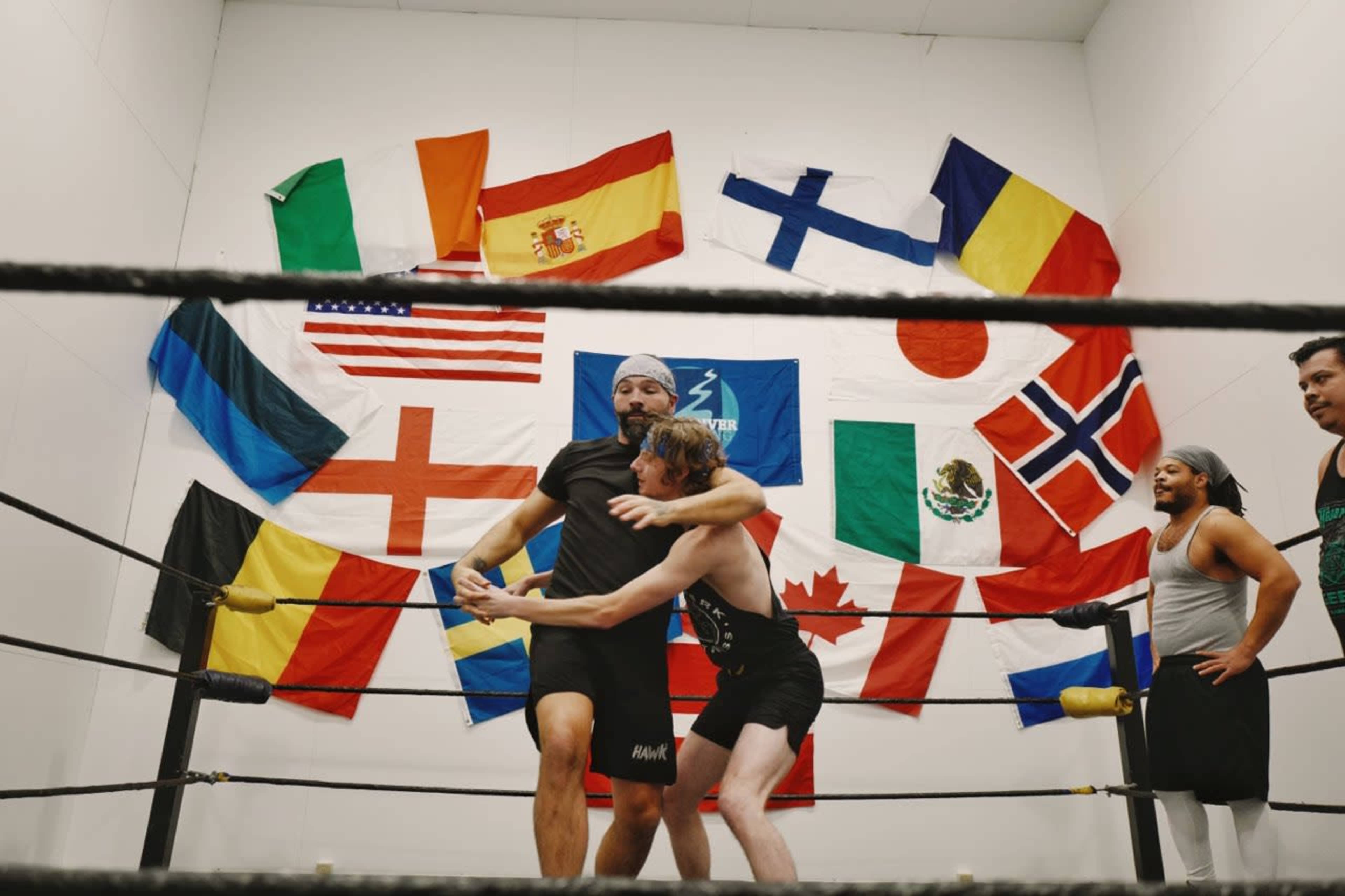 Two wrestlers grapple in a ring surrounded by various national flags hanging on the wall.