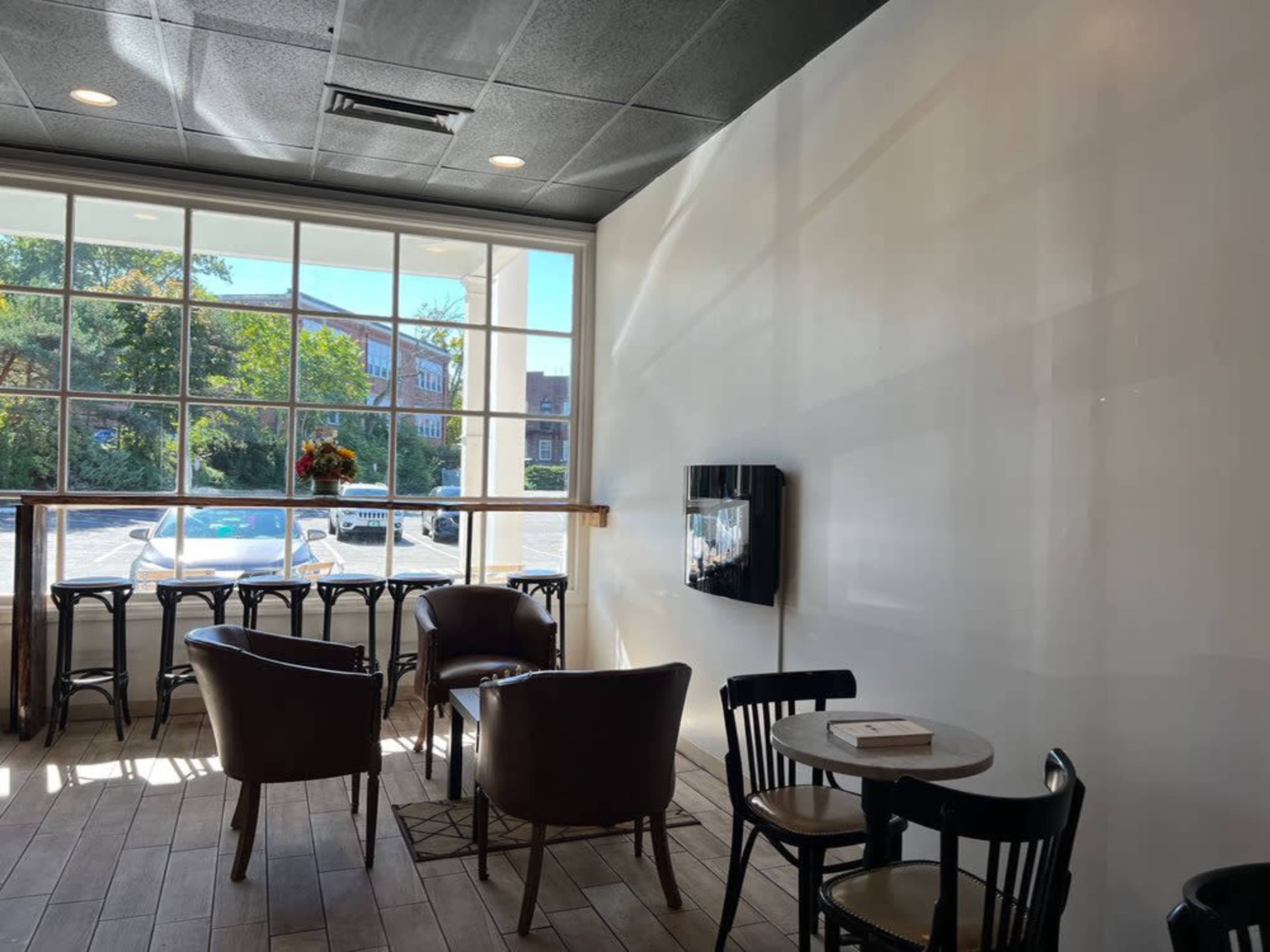 The image shows a brightly lit café interior with a large window overlooking a parking lot, featuring several wooden tables and chairs.
