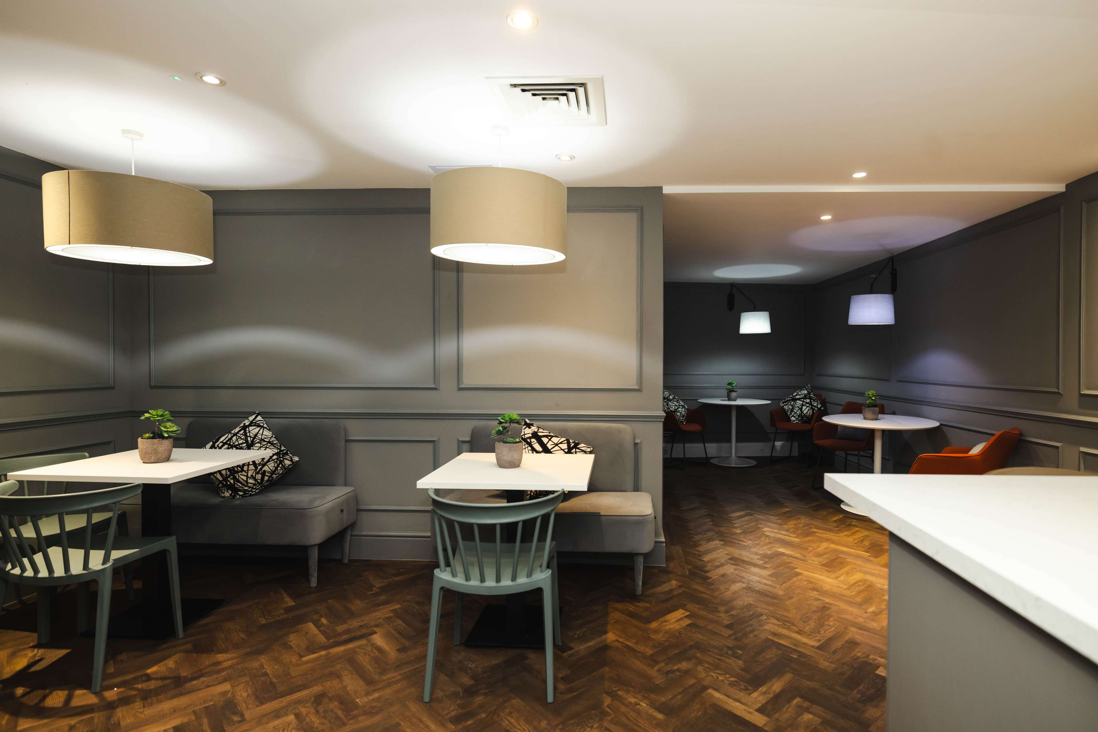 A modern cafe interior with gray walls, wooden flooring, and seating arrangements including tables and chairs, some with decorative plants.