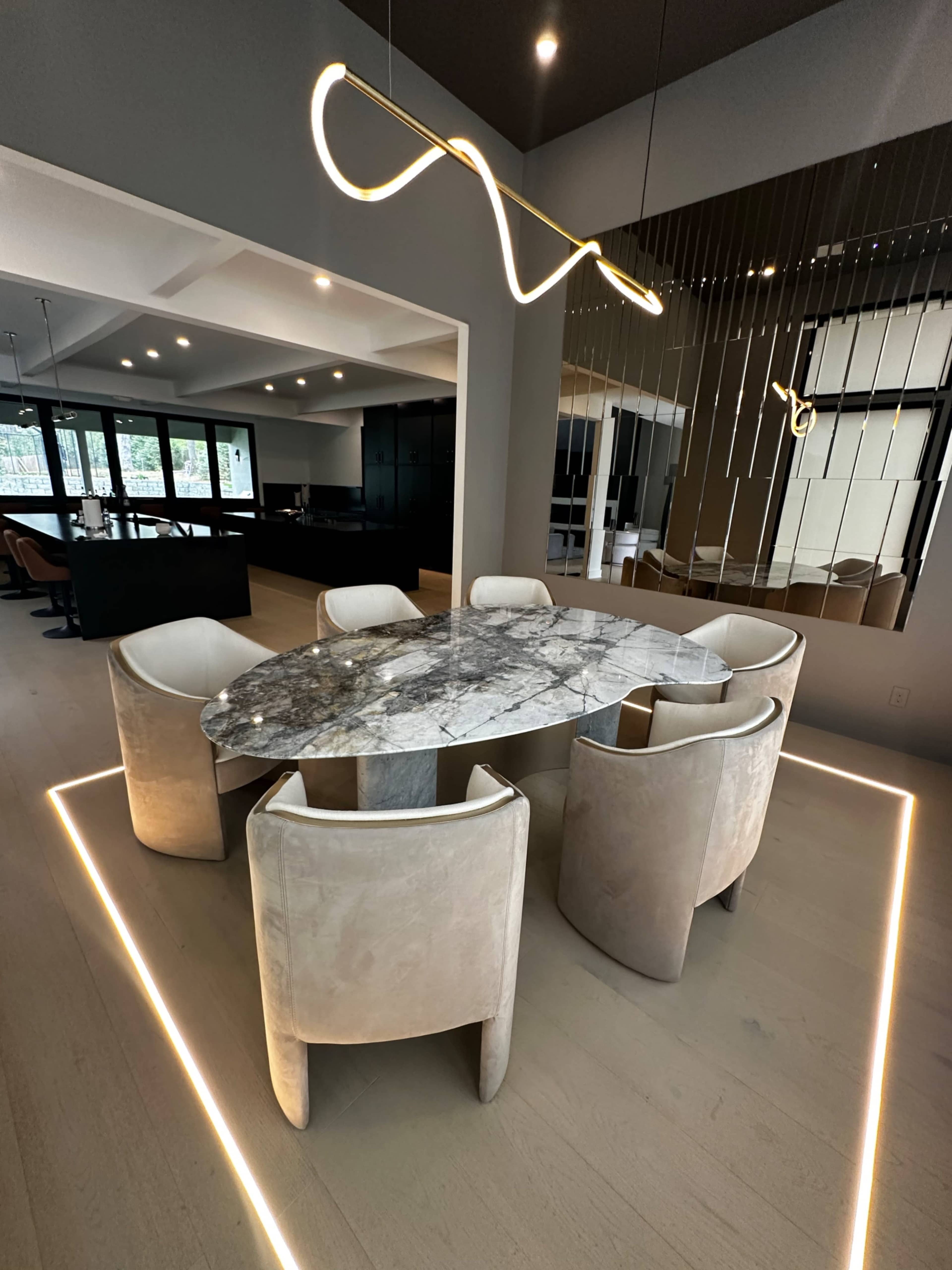 A modern dining area features an oval marble table surrounded by four upholstered chairs, with a sleek light fixture above and a mirrored wall reflecting the space.