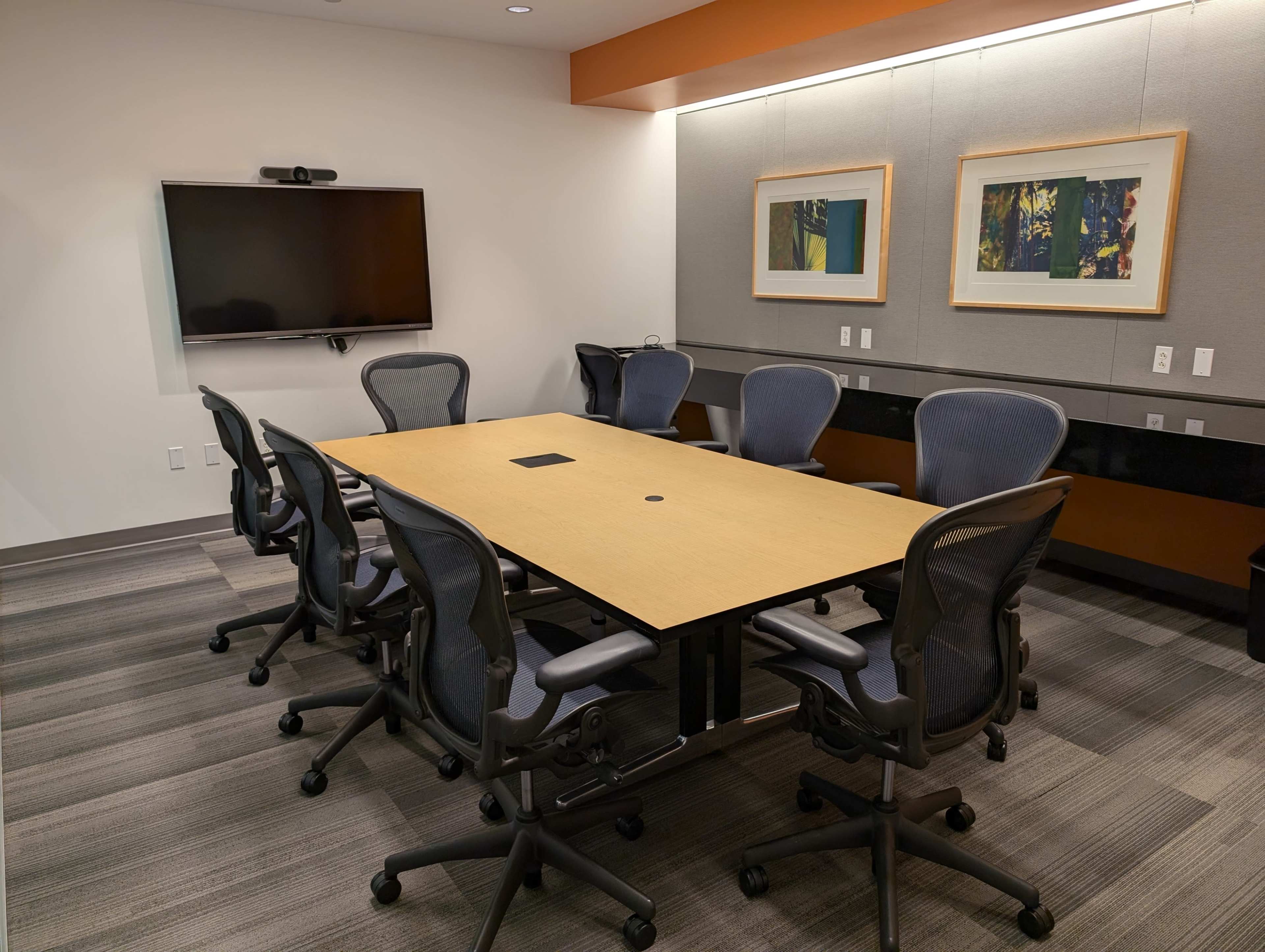 The image shows a modern conference room with a large square table, several ergonomic chairs, a wall-mounted television, and framed artwork.