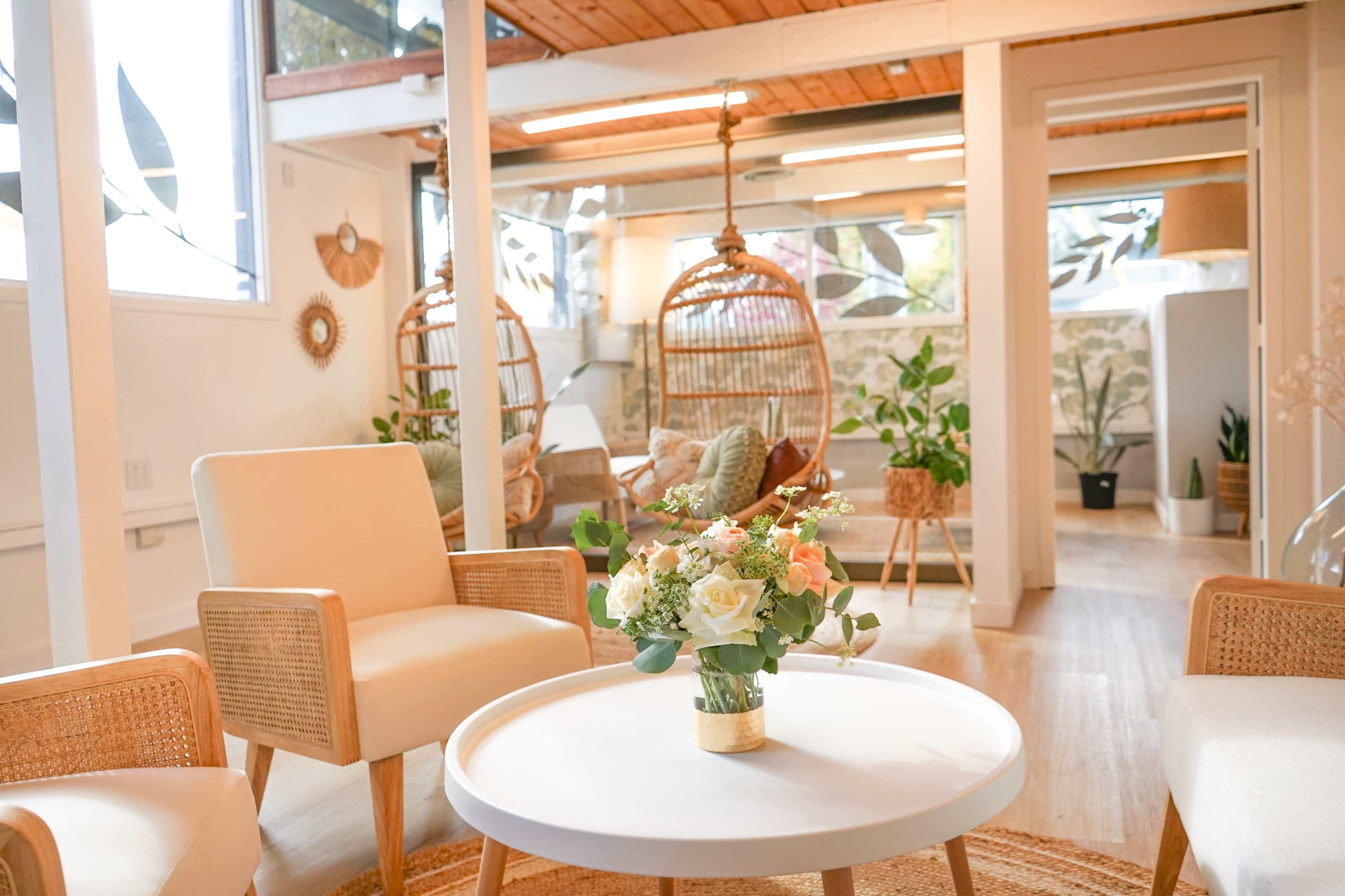 A warmly lit room with two chairs, a round table adorned with a floral arrangement, and hanging wicker chairs in the background.