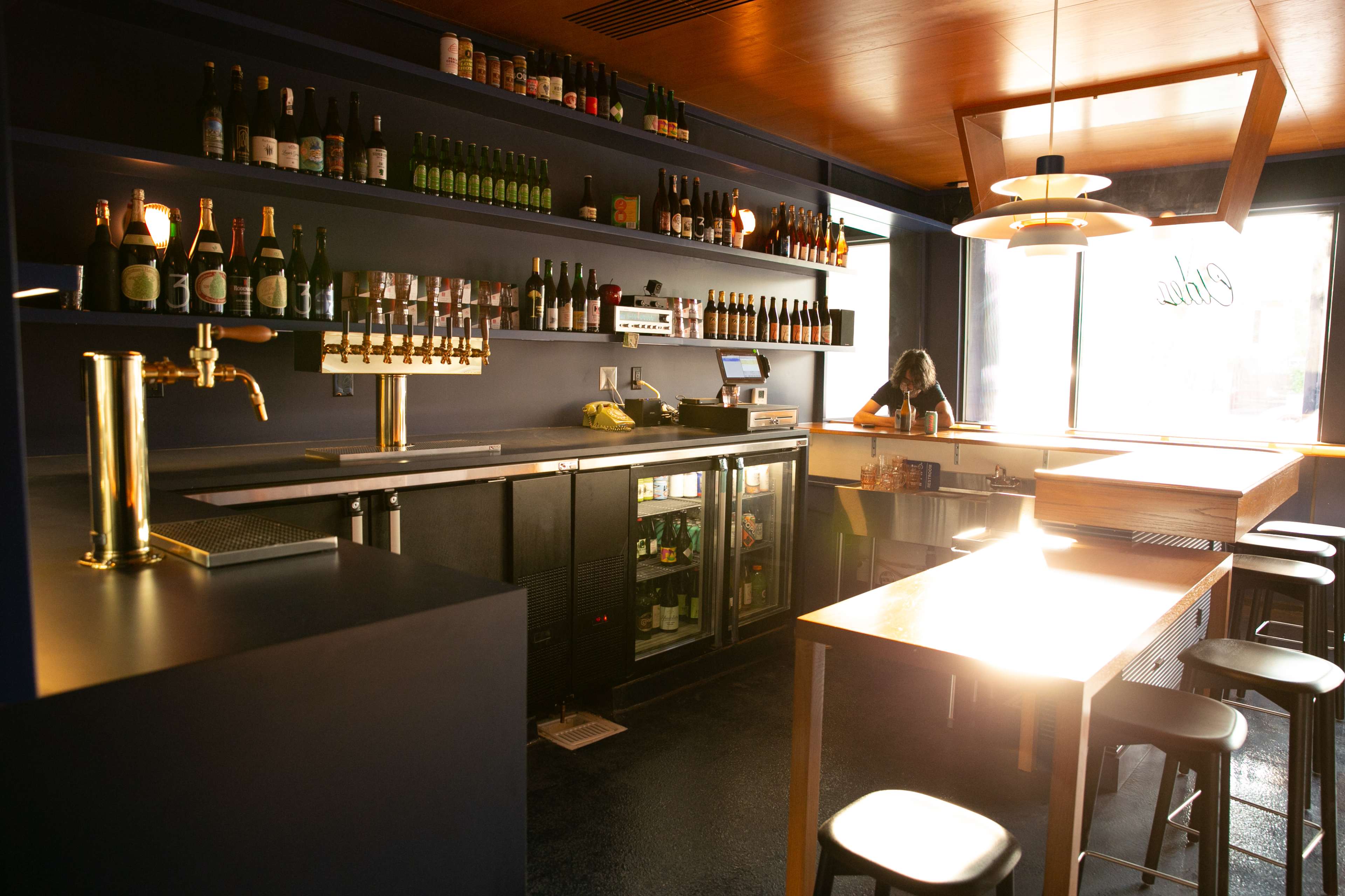 A modern bar area with a counter, shelves stocked with bottled drinks, and a bartender preparing in the background.