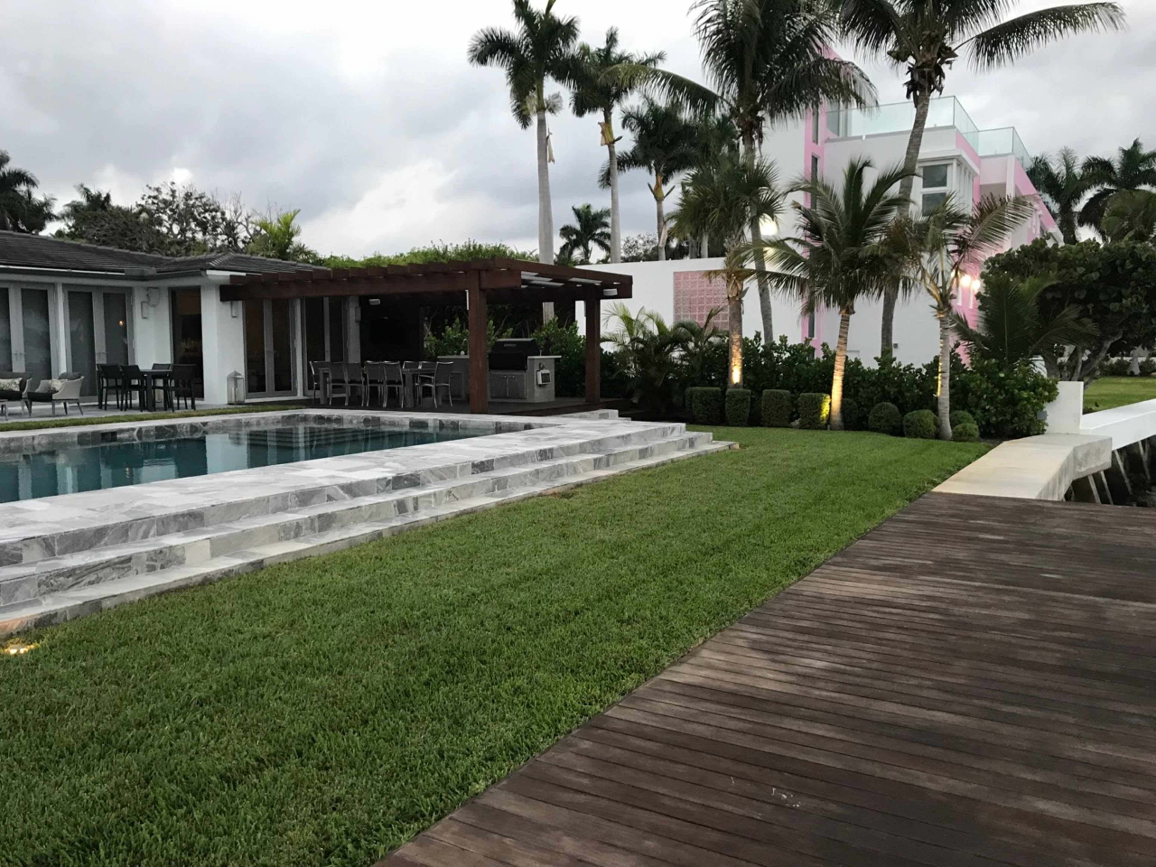 The image shows a modern home with a swimming pool, surrounded by manicured lawns and palm trees, along with a wooden walkway leading to the water.