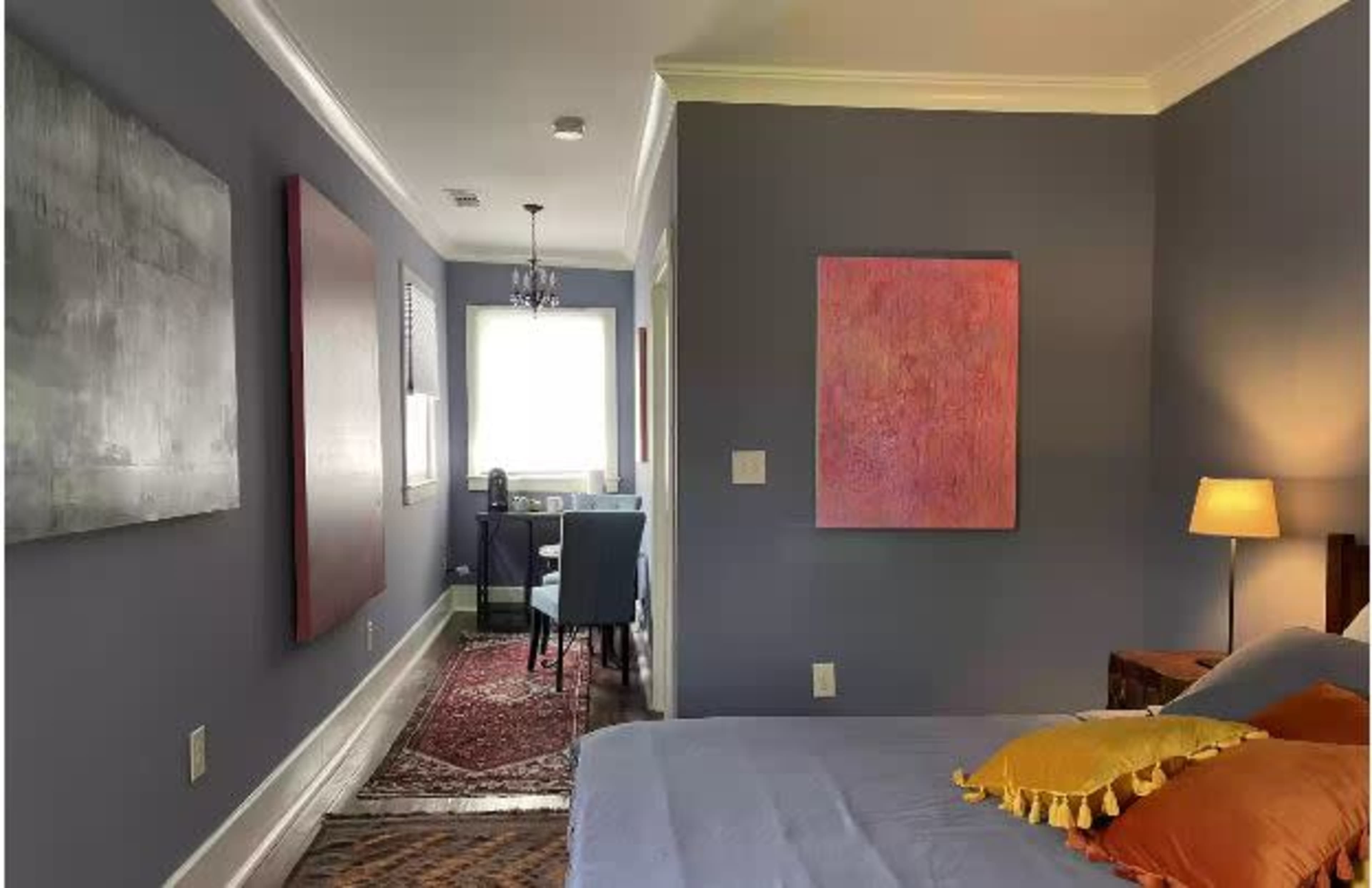 The image shows a bedroom with gray walls, a bed with colorful pillows, a lamp, and a small desk and chair by a window.