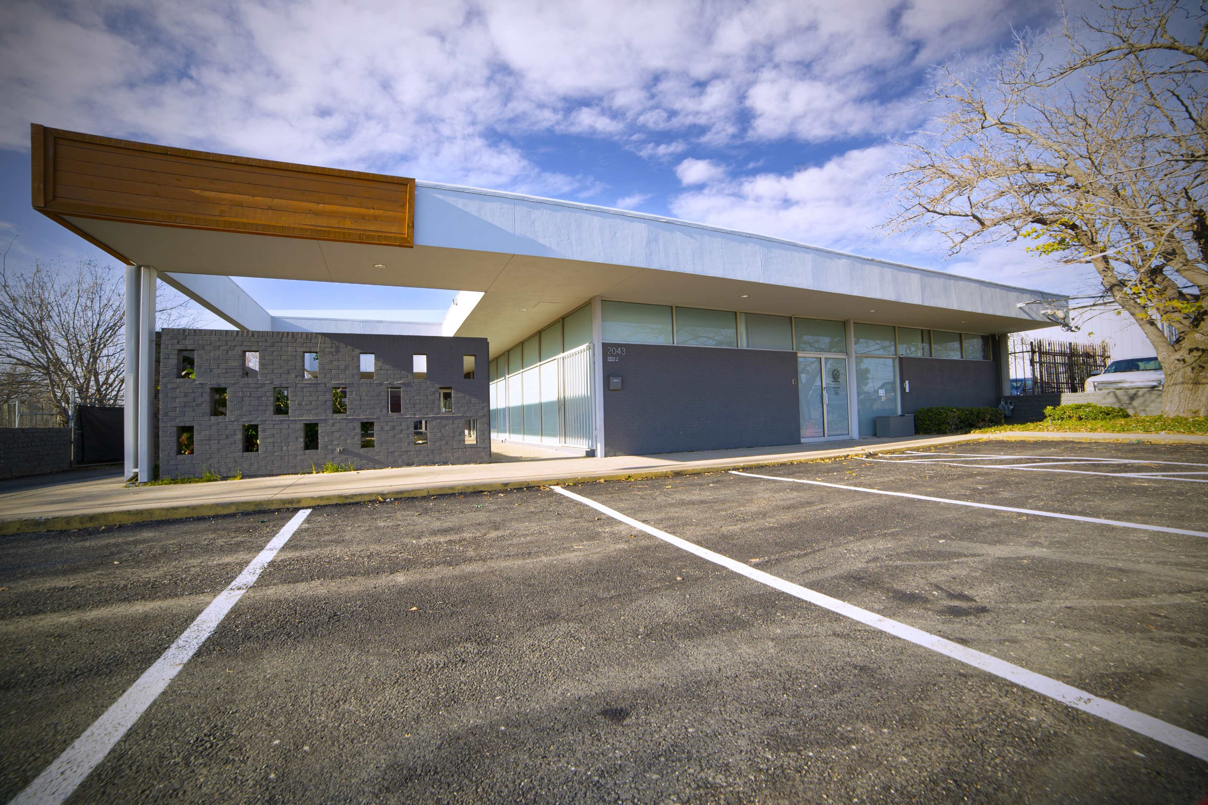 A modern building with large windows, a wooden overhang, and a distinctive wall design is situated in an expansive, empty parking lot.