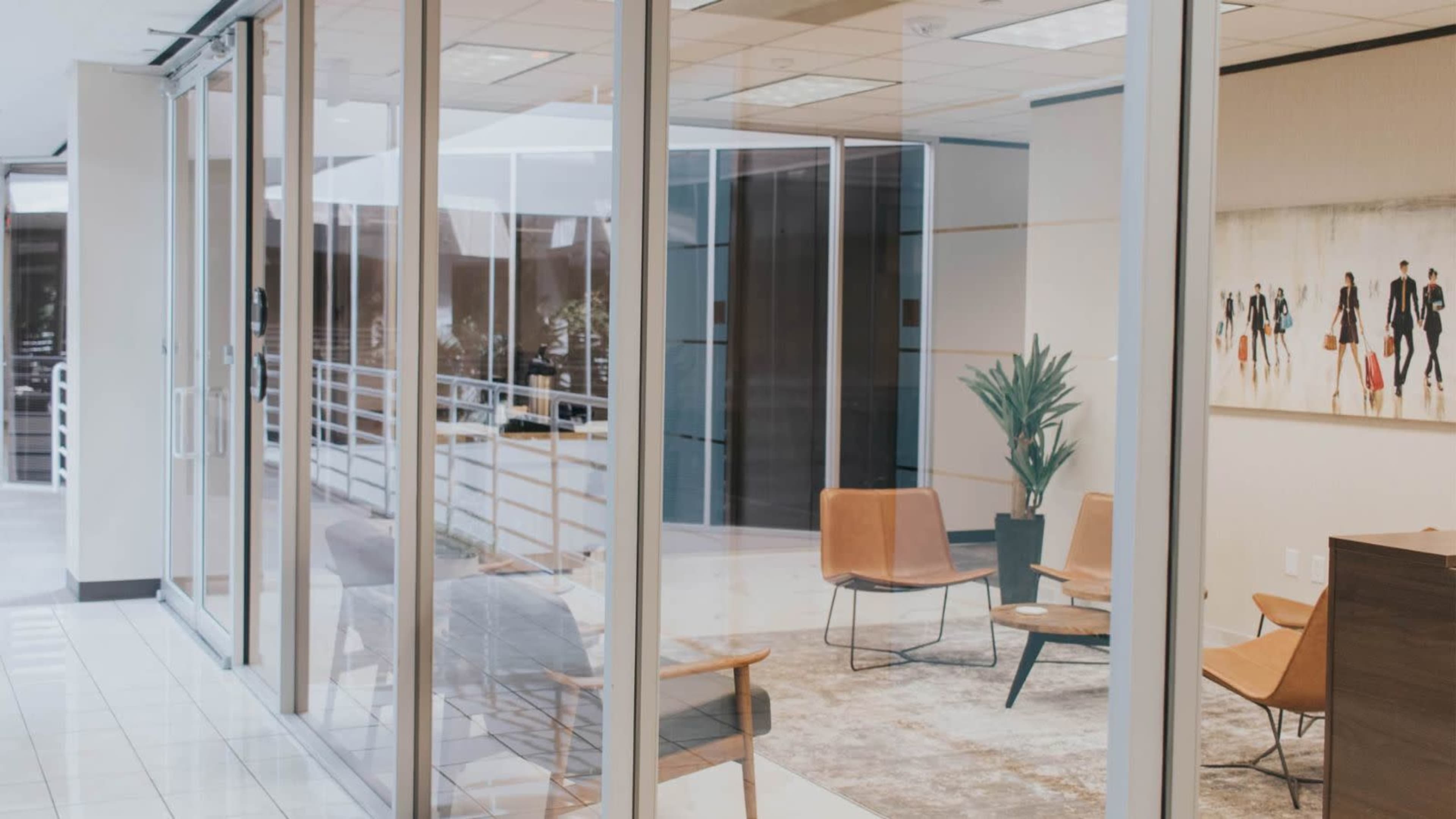 The image shows a modern office interior with glass walls, featuring a seating area with four chairs, a small table, and a decorative plant.