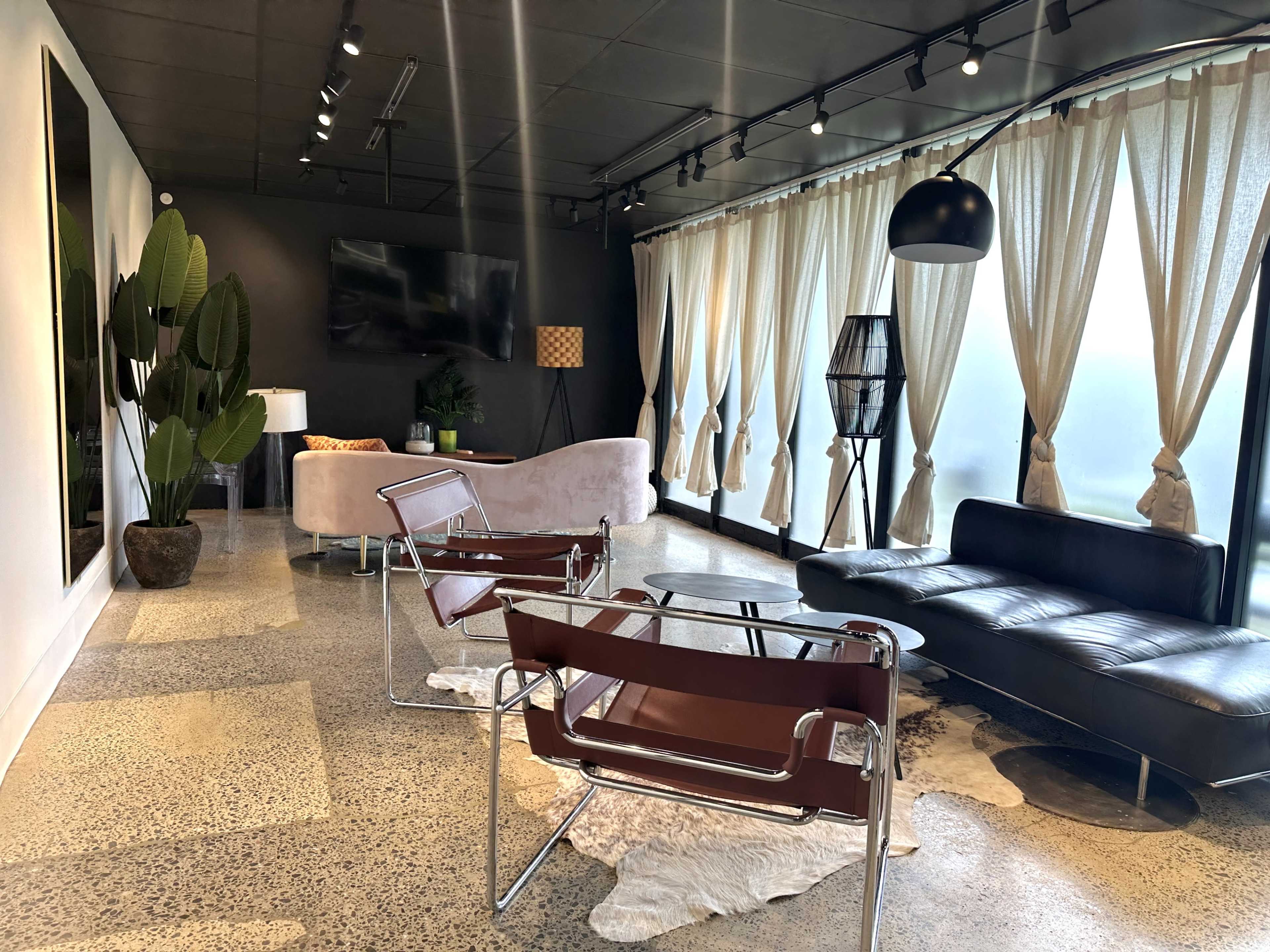 The image shows a modern interior space featuring a pink sofa, a black leather couch, metallic chairs, large windows with sheer curtains, and decorative plants.