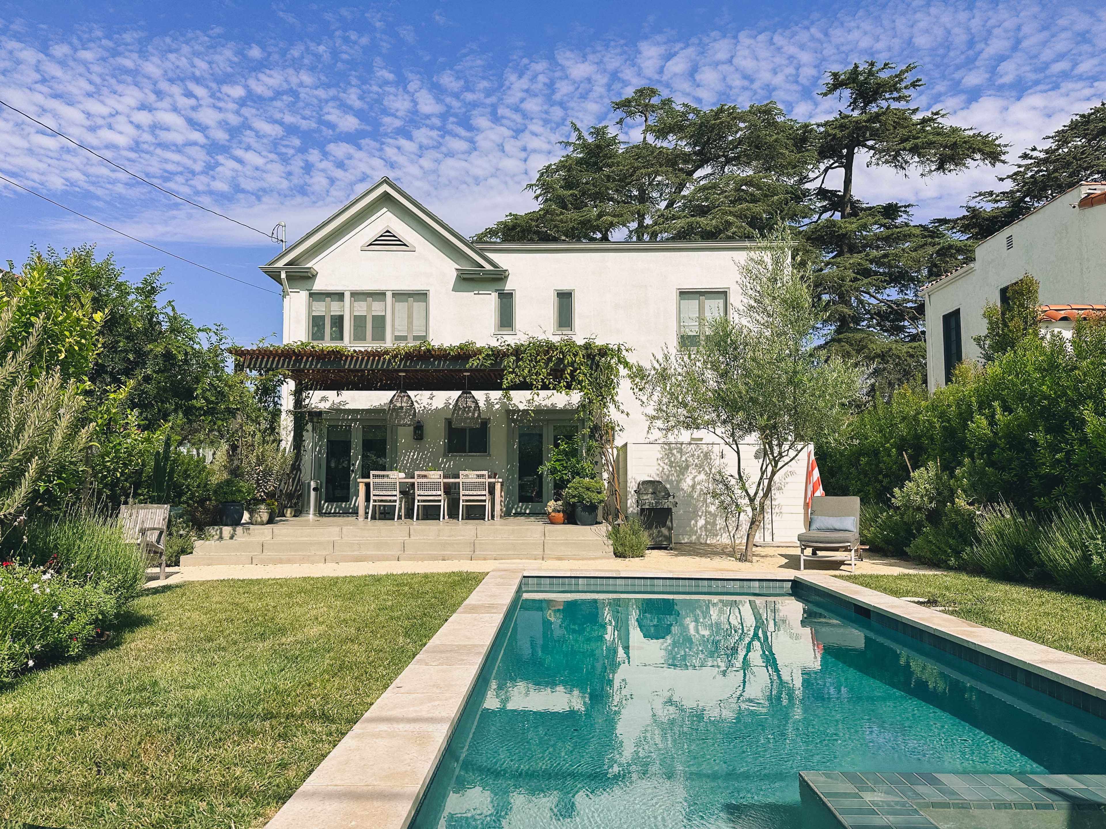 A two-story white house with a green lawn and a swimming pool, surrounded by lush greenery under a blue sky.