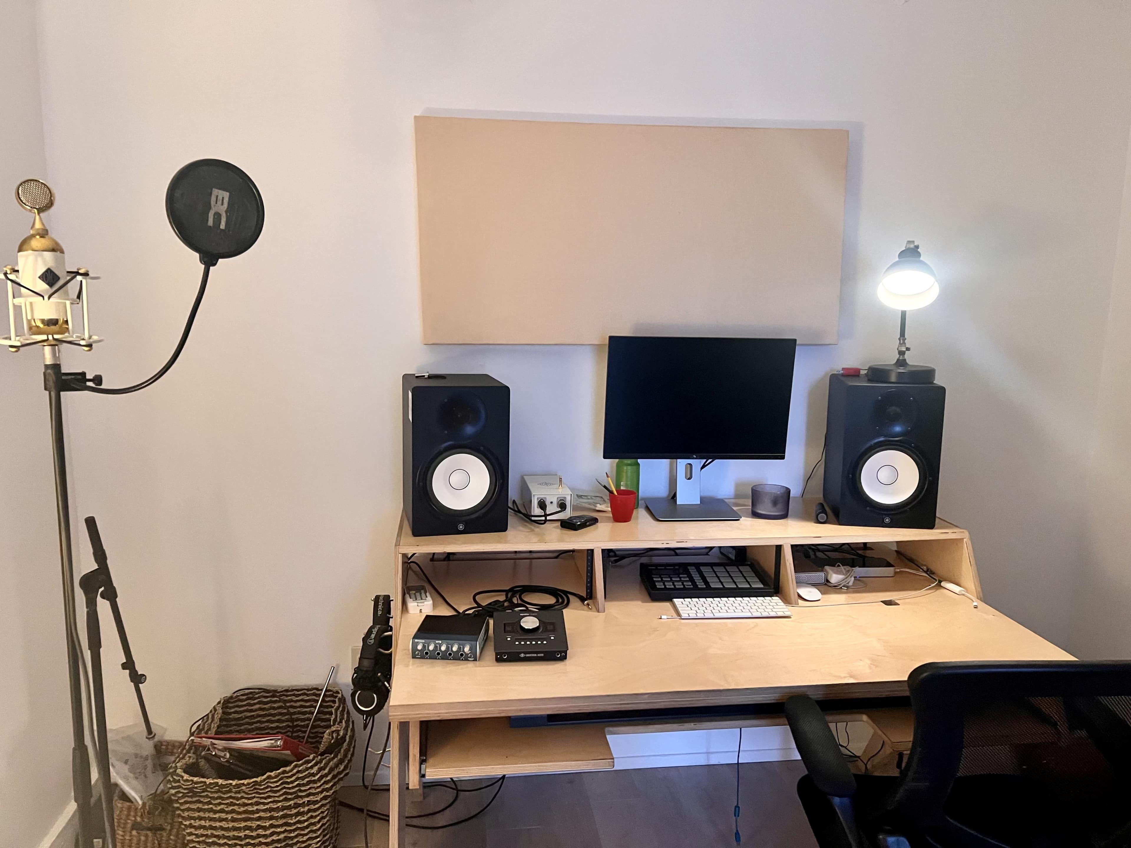 The image shows a recording studio setup featuring a desktop computer, two studio monitors, a microphone on a stand, and various audio equipment arranged on a wooden desk.