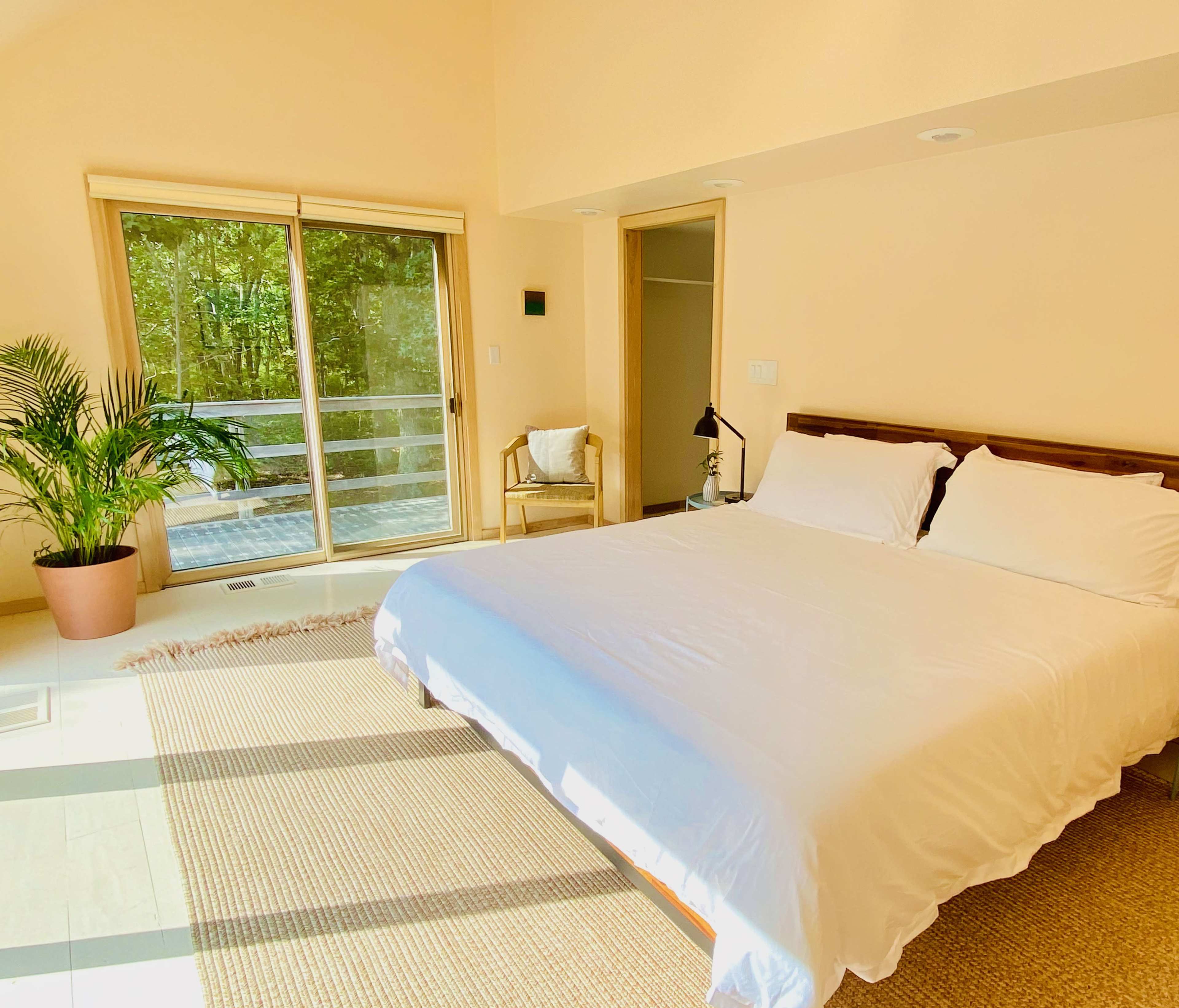 The image shows a minimalist bedroom with a large bed, a chair, and sliding glass doors leading to a deck surrounded by greenery.