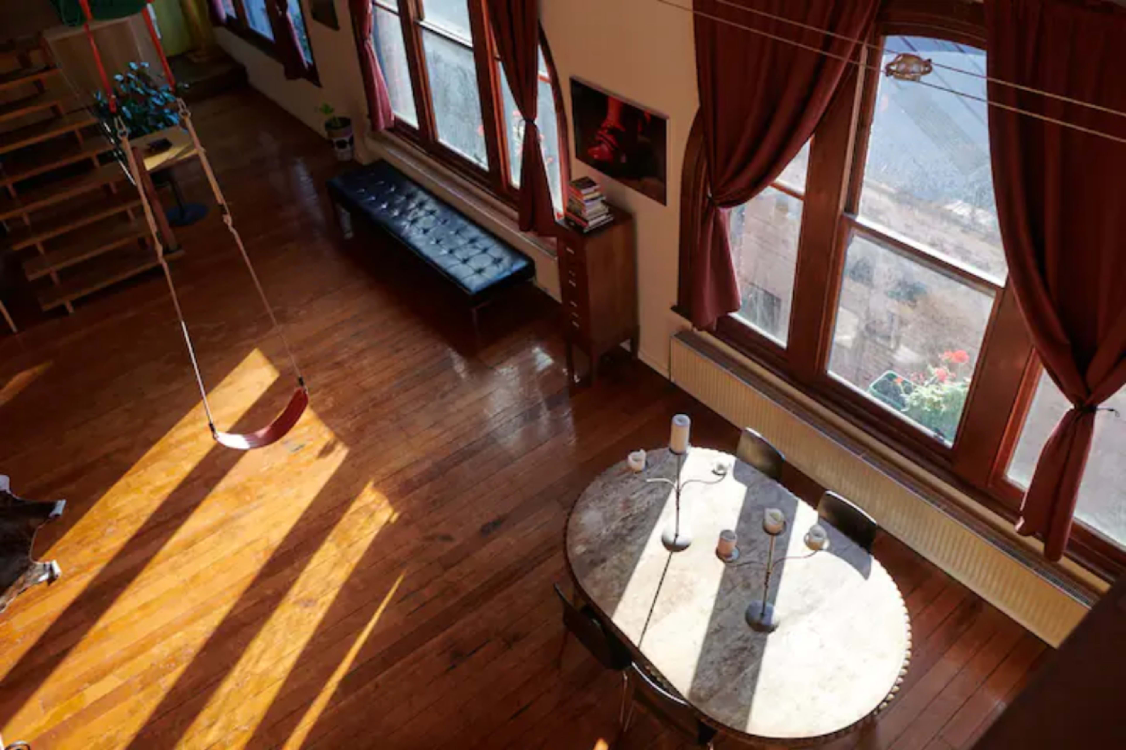 A spacious, well-lit room with wooden floors, large windows, a round dining table, and a swing hanging from the ceiling.