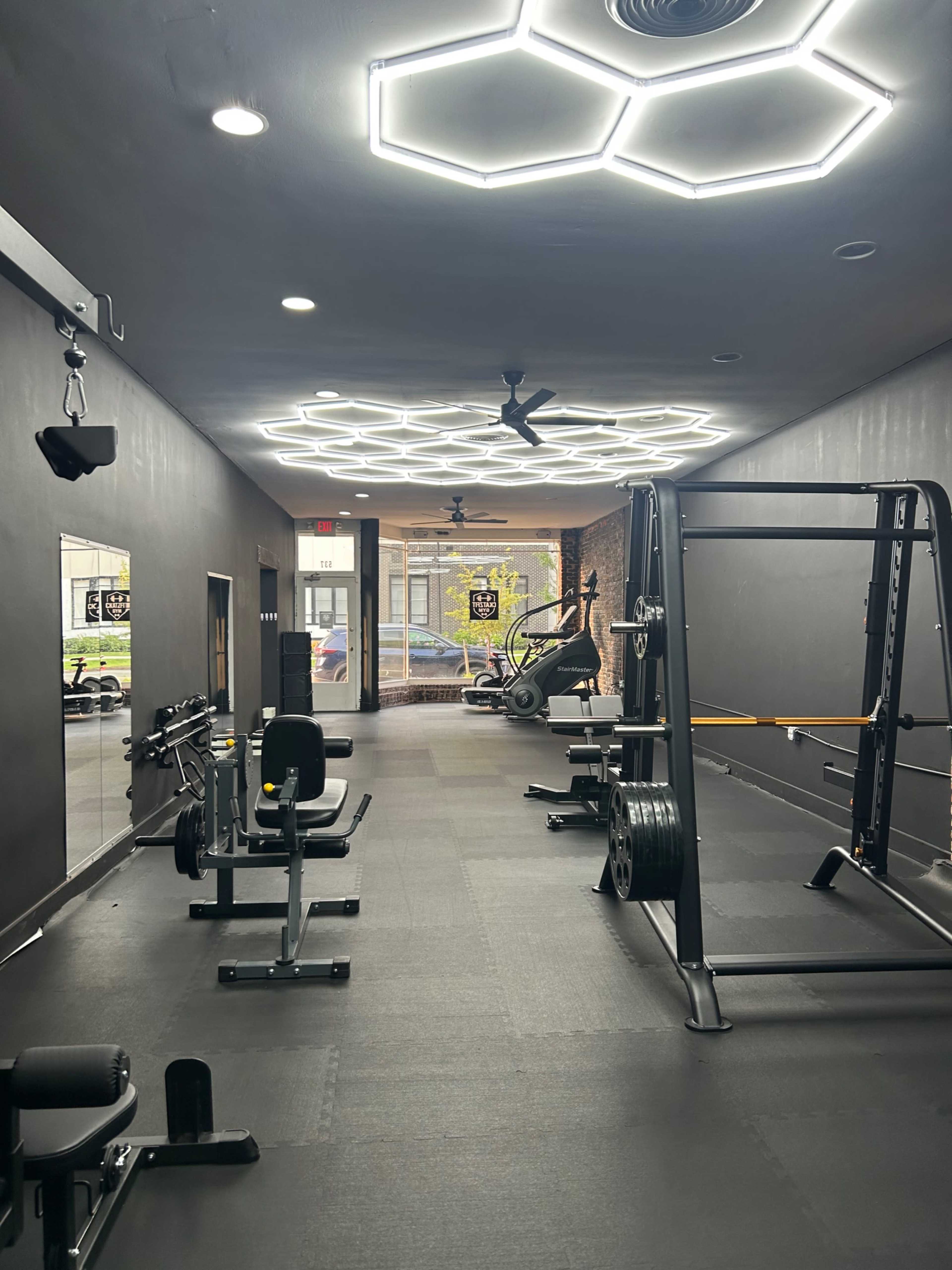The image shows a modern gym interior with hexagonal lighting, gym equipment on black flooring, and large windows allowing natural light to enter.