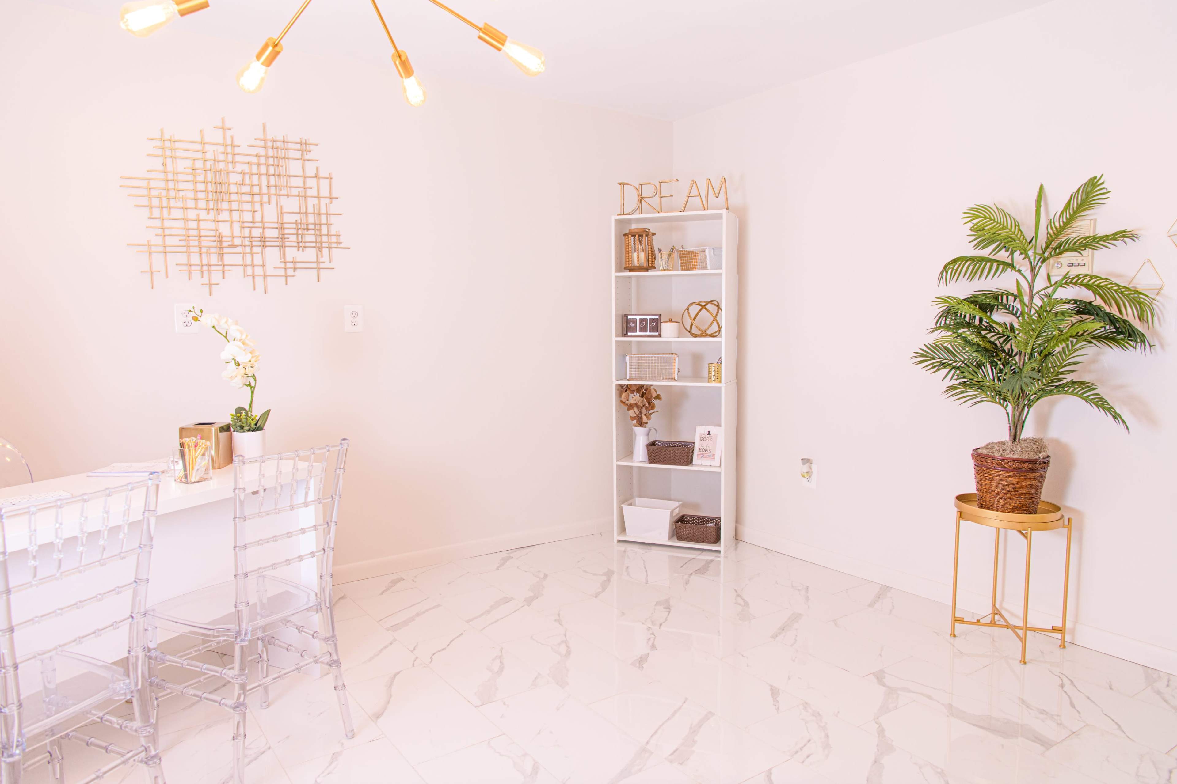 A modern, minimalistic room features a glass-top dining table with transparent chairs, a white bookshelf filled with decorative items, and a potted plant placed on a gold stand.