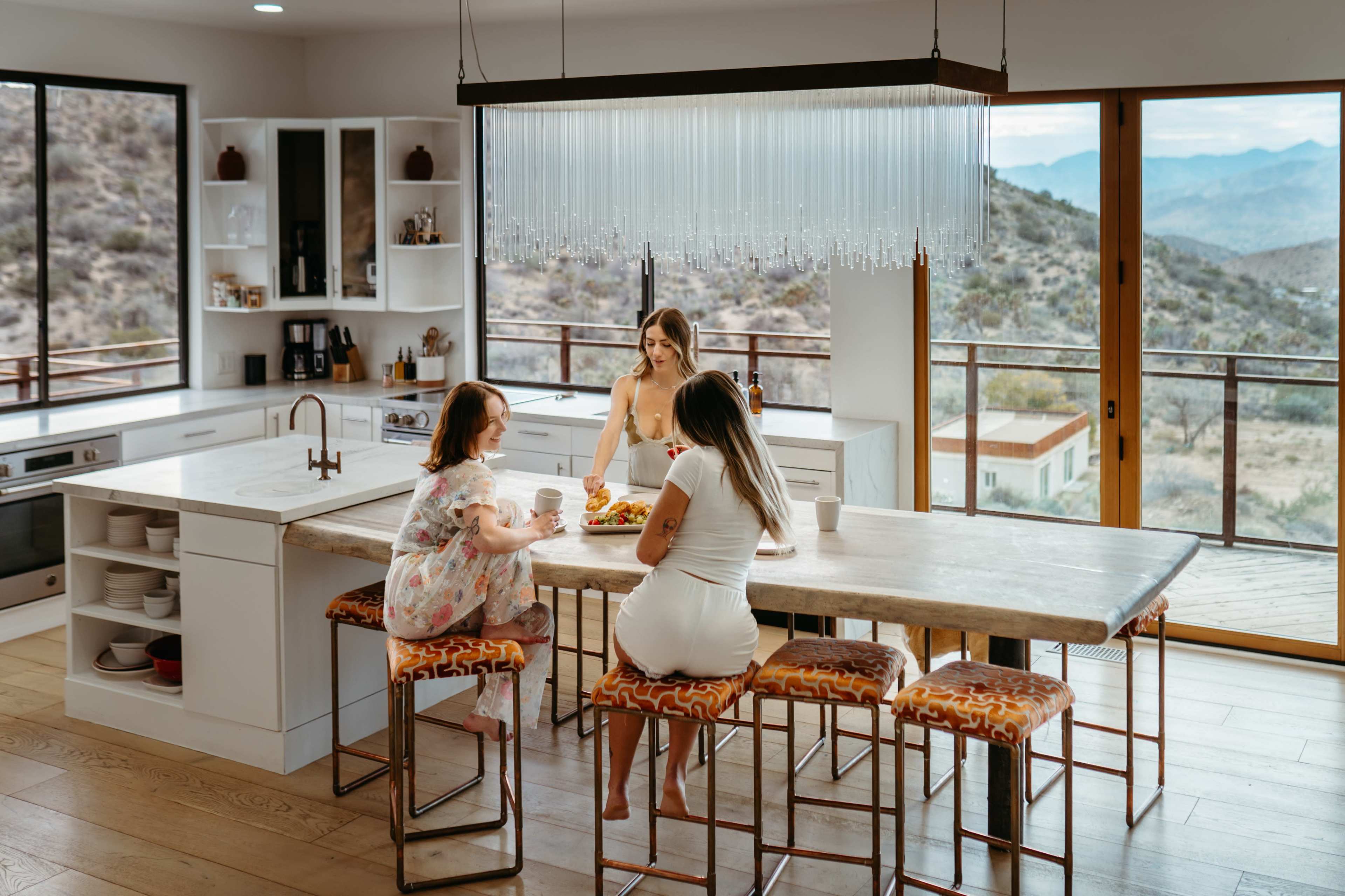 Three women sit at a large kitchen island in a modern kitchen, with scenic mountain views visible through the large windows.