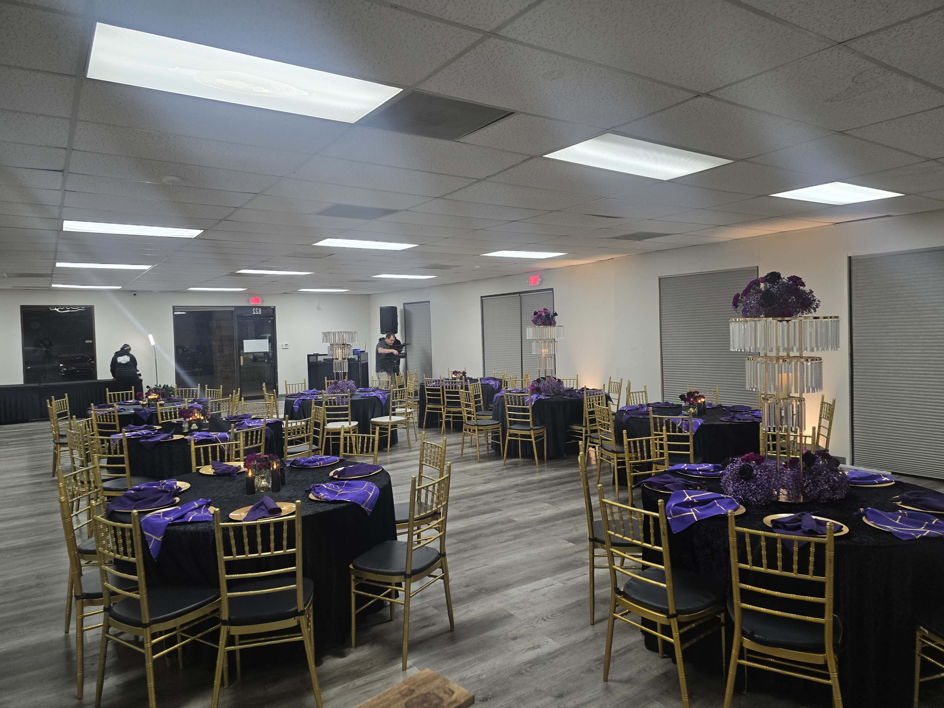 The image shows a banquet hall set up with round tables covered in black tablecloths and adorned with purple decorations, surrounded by gold chairs.