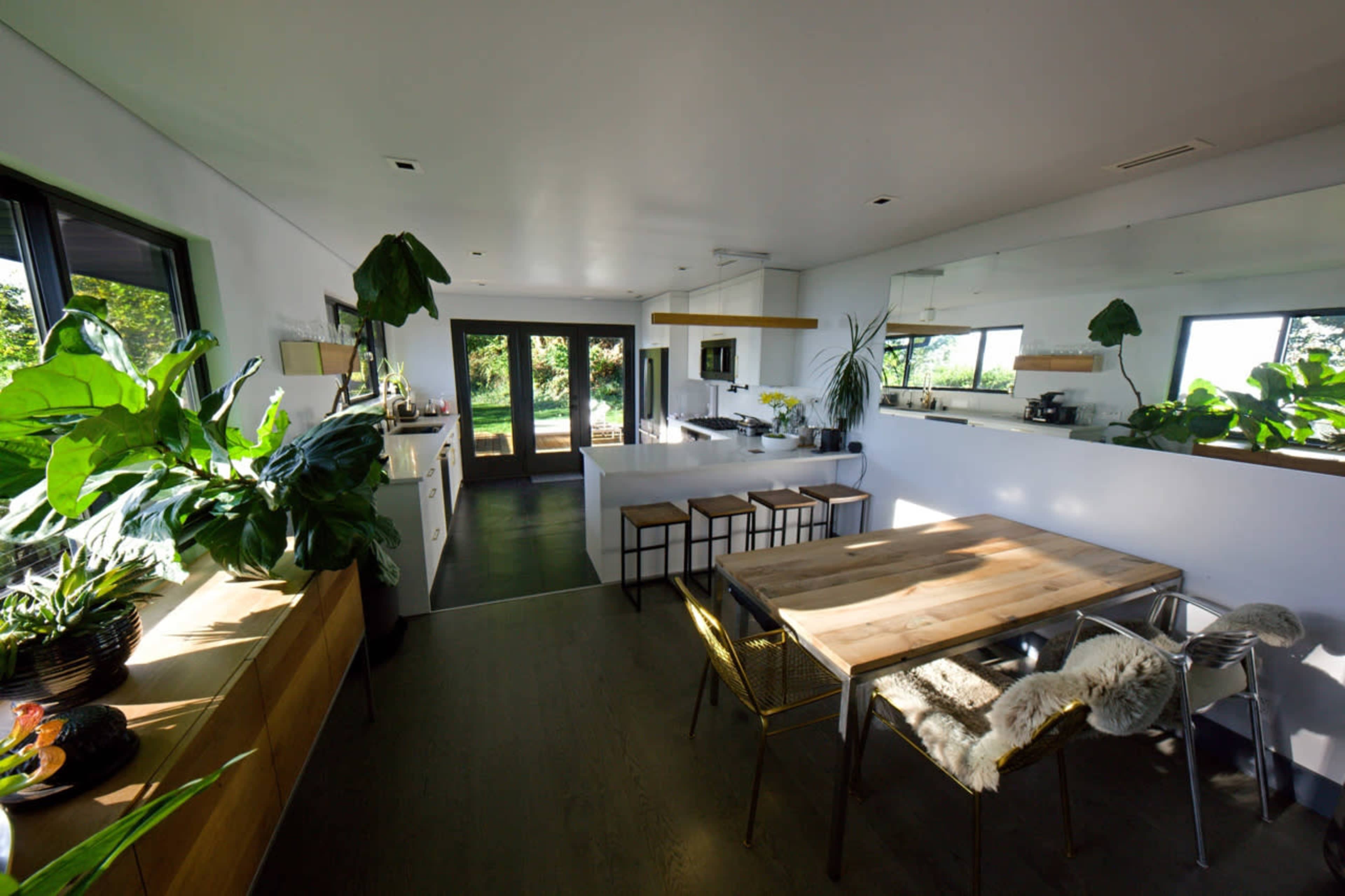 A modern kitchen and dining area with large windows, a wooden table, metal chairs, and green plants.