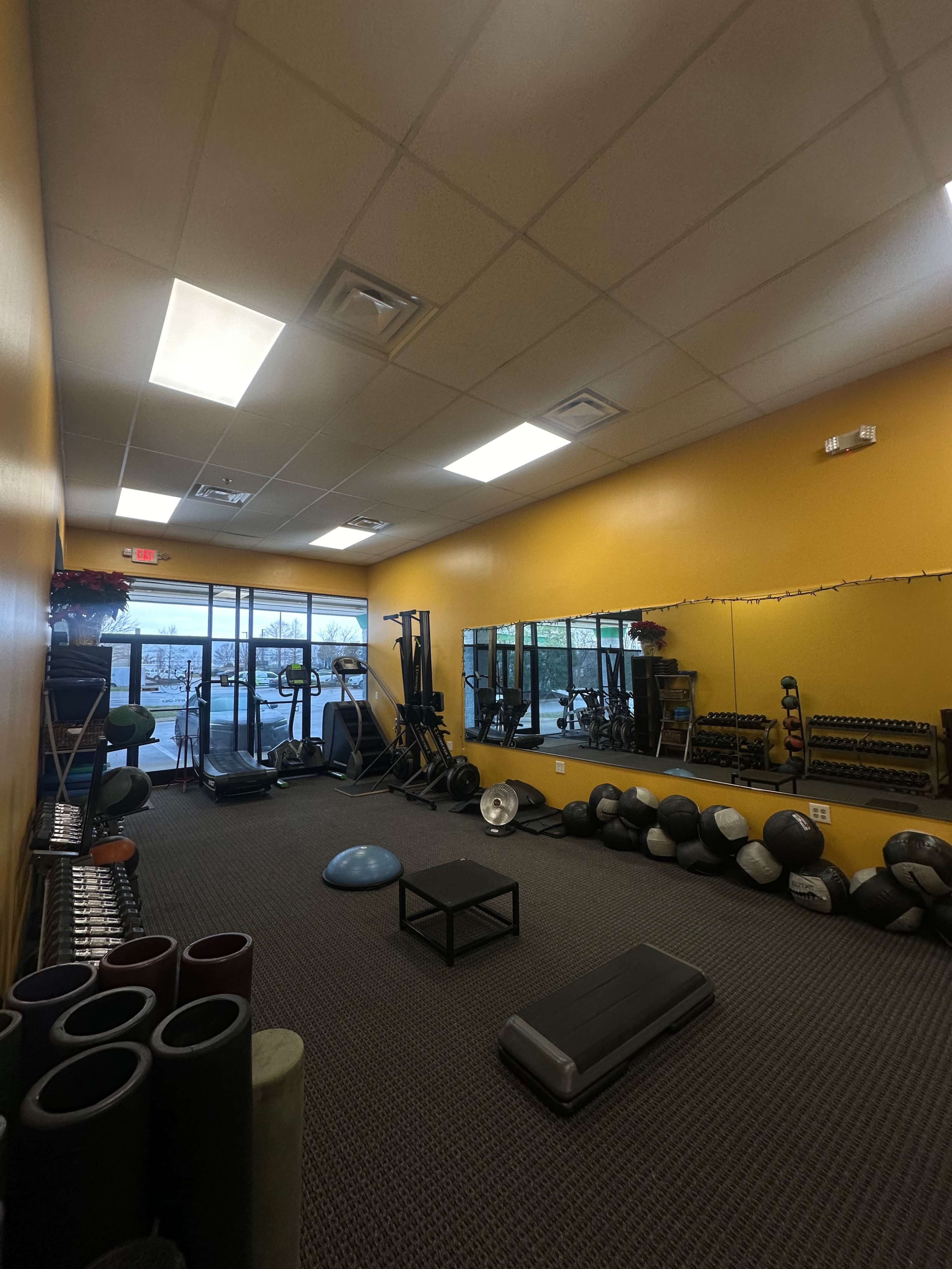 The image shows a fitness studio with exercise equipment, including weights and mats, alongside large windows allowing natural light.