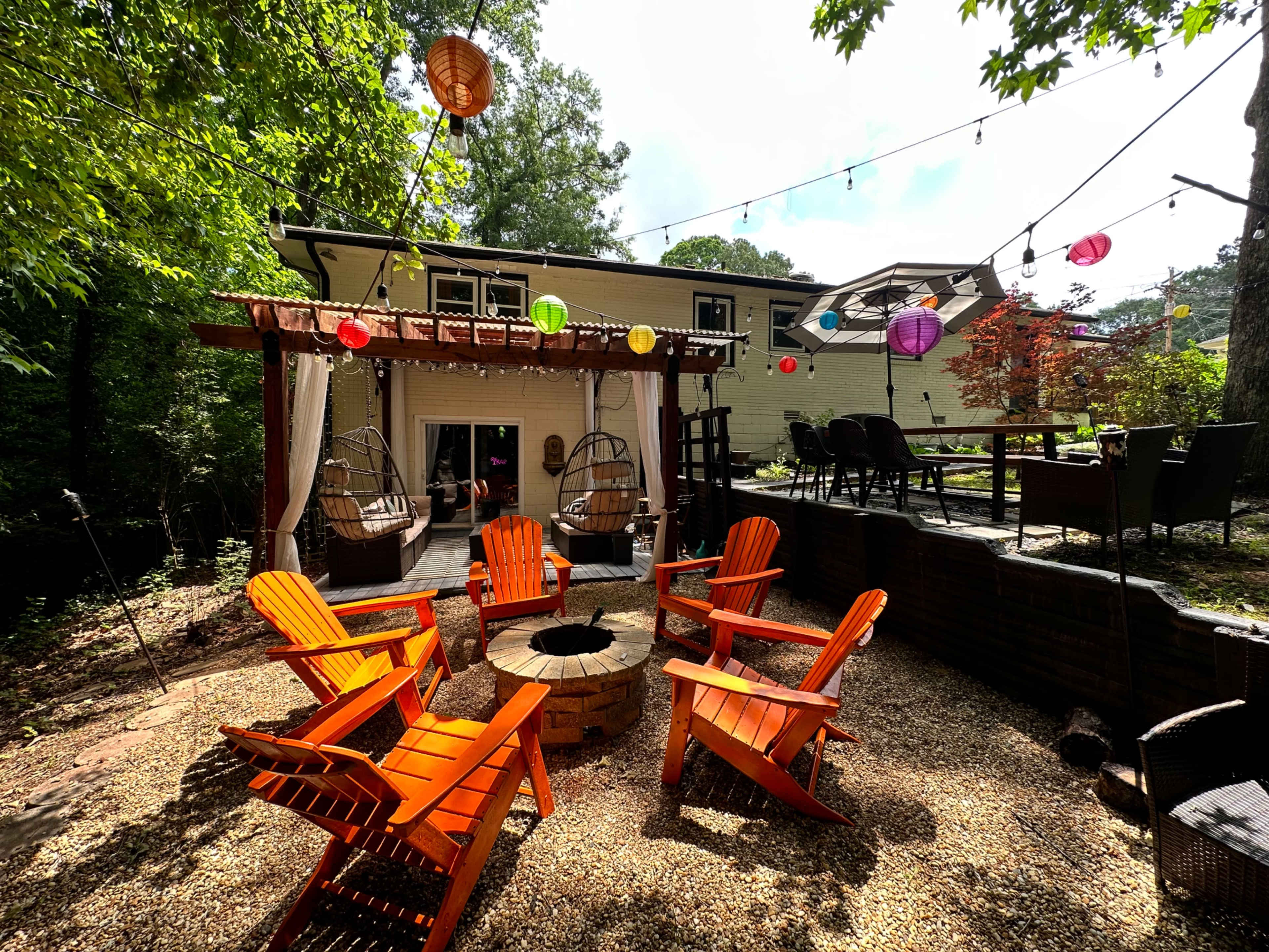 The scene shows a backyard with a stone fire pit surrounded by orange Adirondack chairs, hanging lanterns, and a patio area with a dining table.