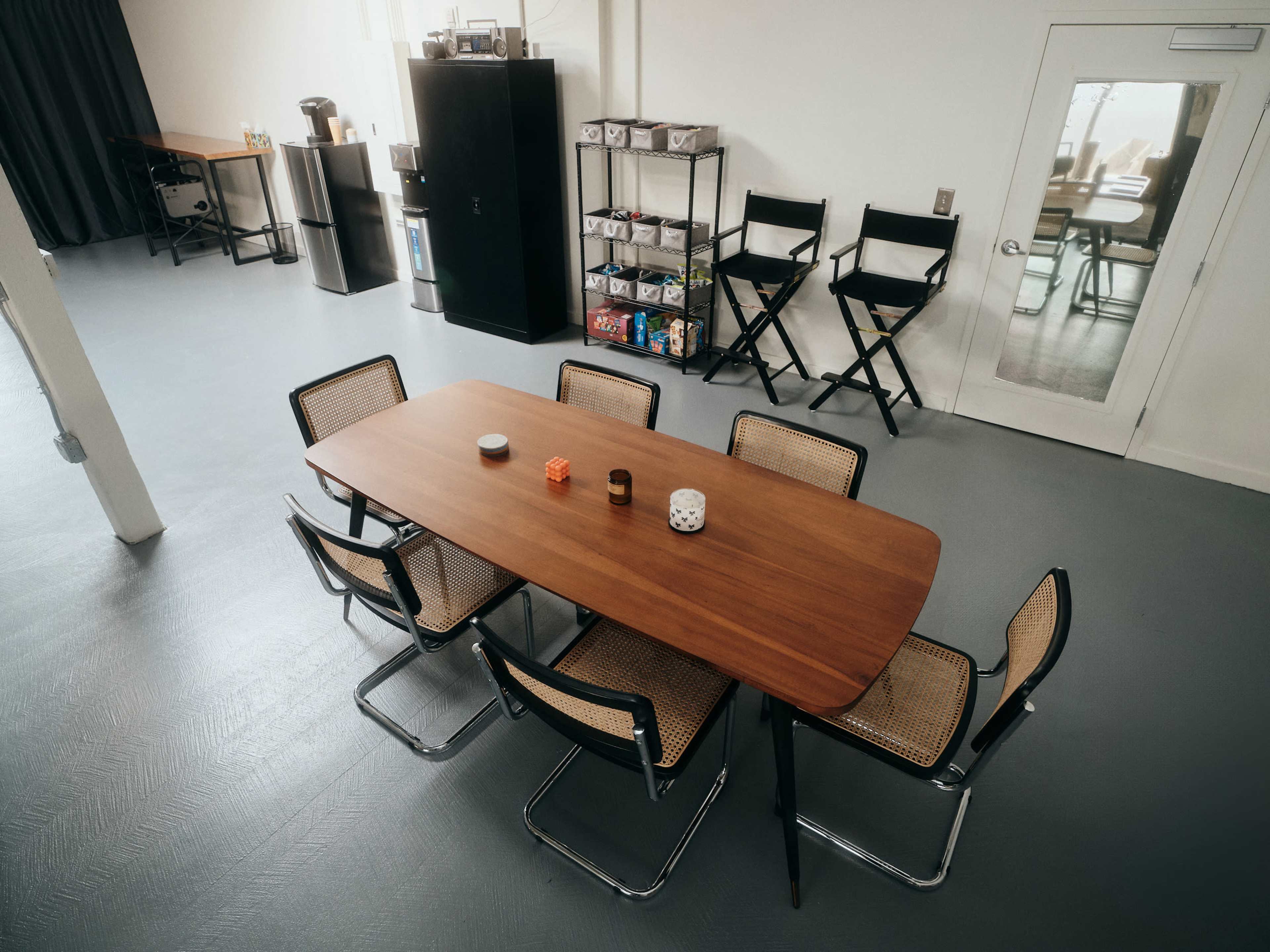 A modern meeting room features a large wooden table surrounded by six chairs, with a kitchenette and storage shelves visible in the background.