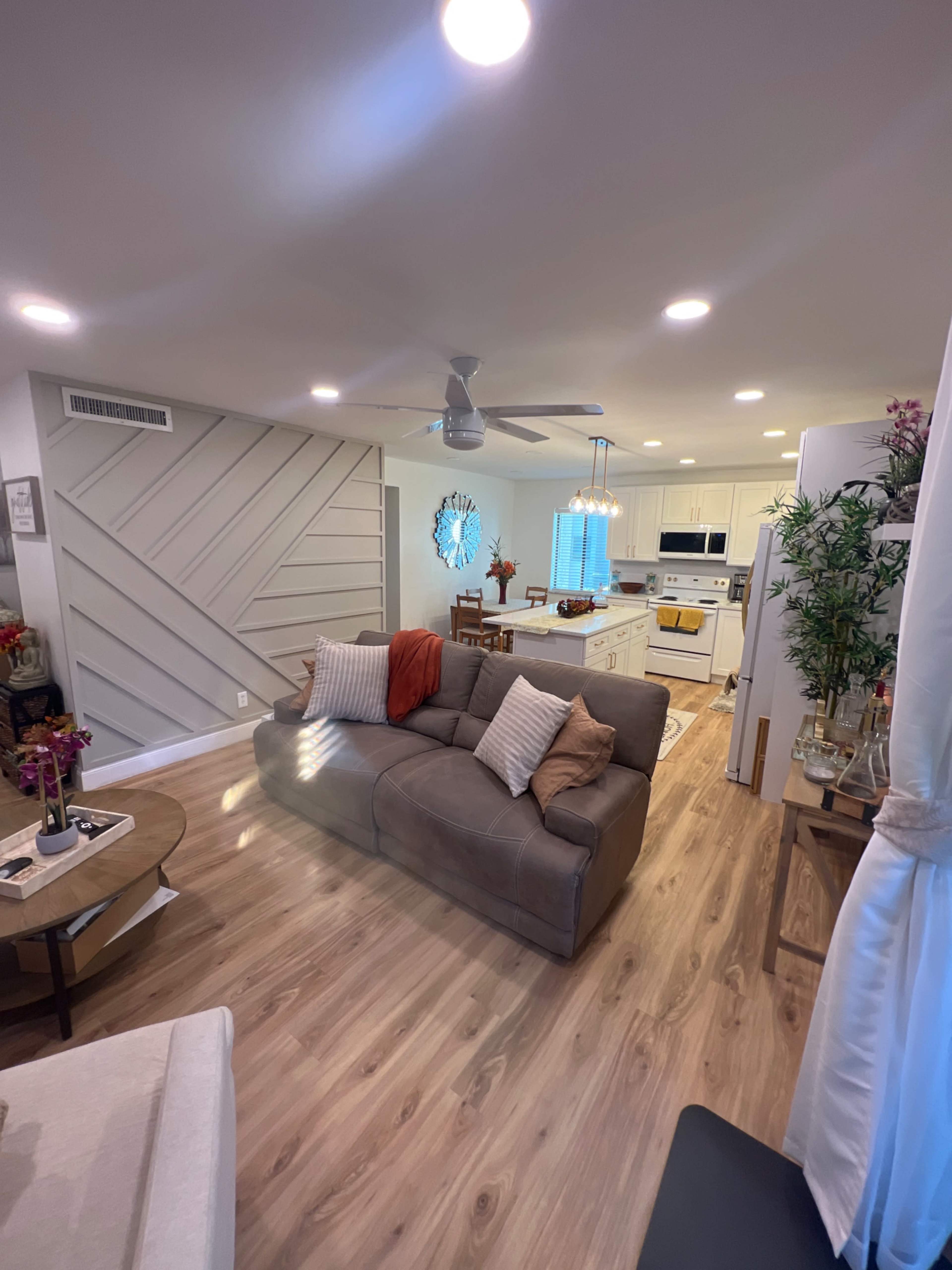 The image shows a modern living room with a light-colored sofa, a coffee table, and a view into a kitchen area, featuring wood flooring and bright lighting.