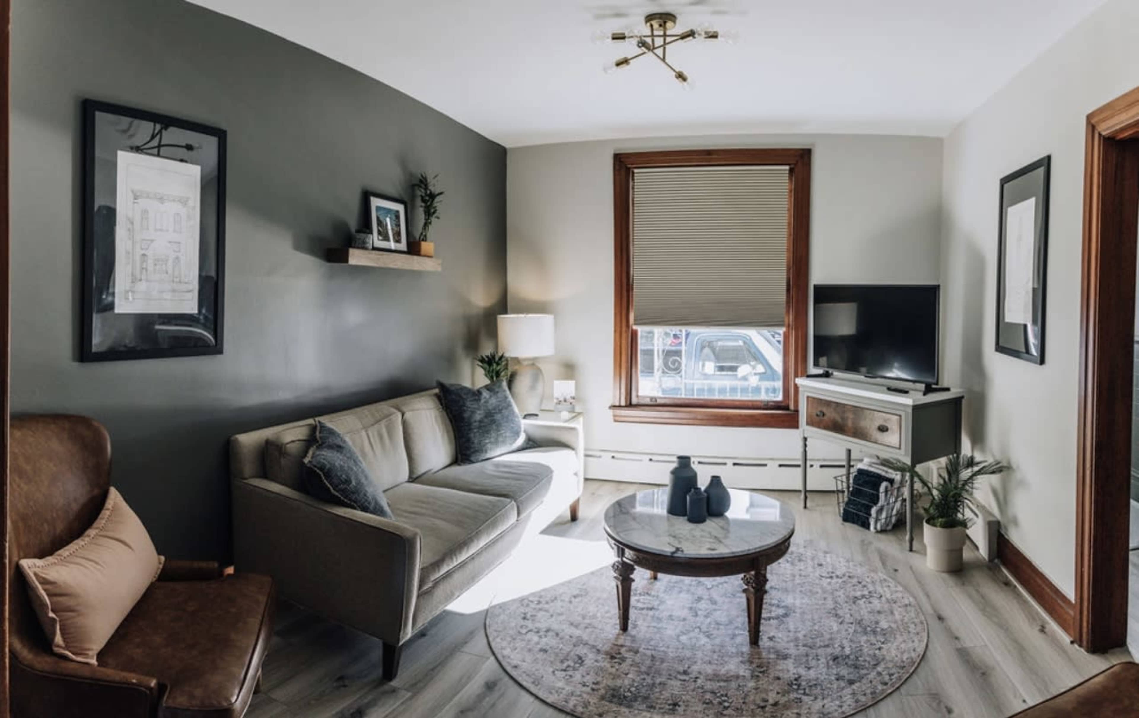 A cozy living room with a gray accent wall, a modern sofa, a round coffee table, and a television on a wooden stand, illuminated by natural light from a window.