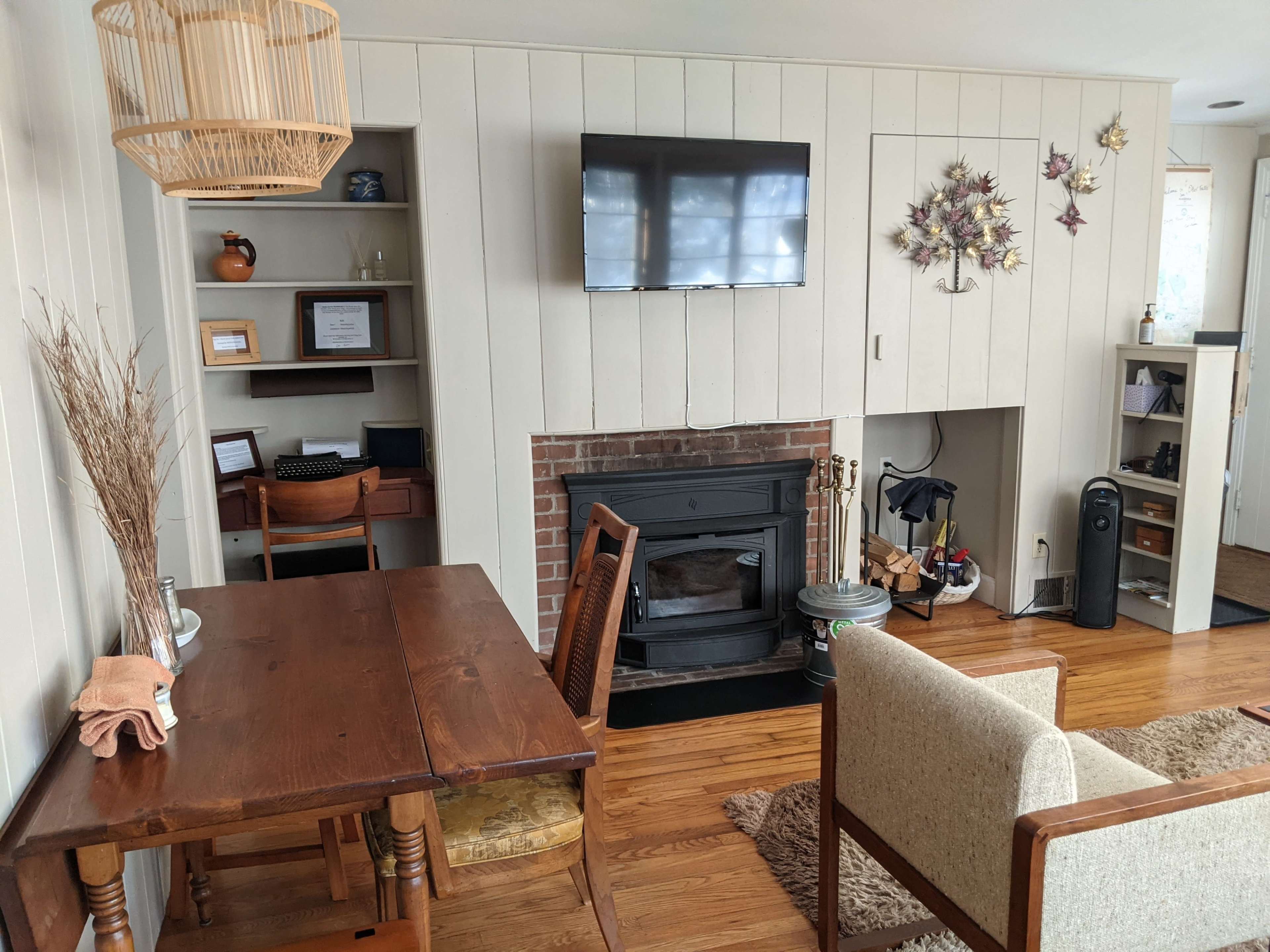 A cozy living space featuring a wooden dining table, two chairs, a fireplace, a television mounted on the wall, and decorative elements like dried plants and wall art.