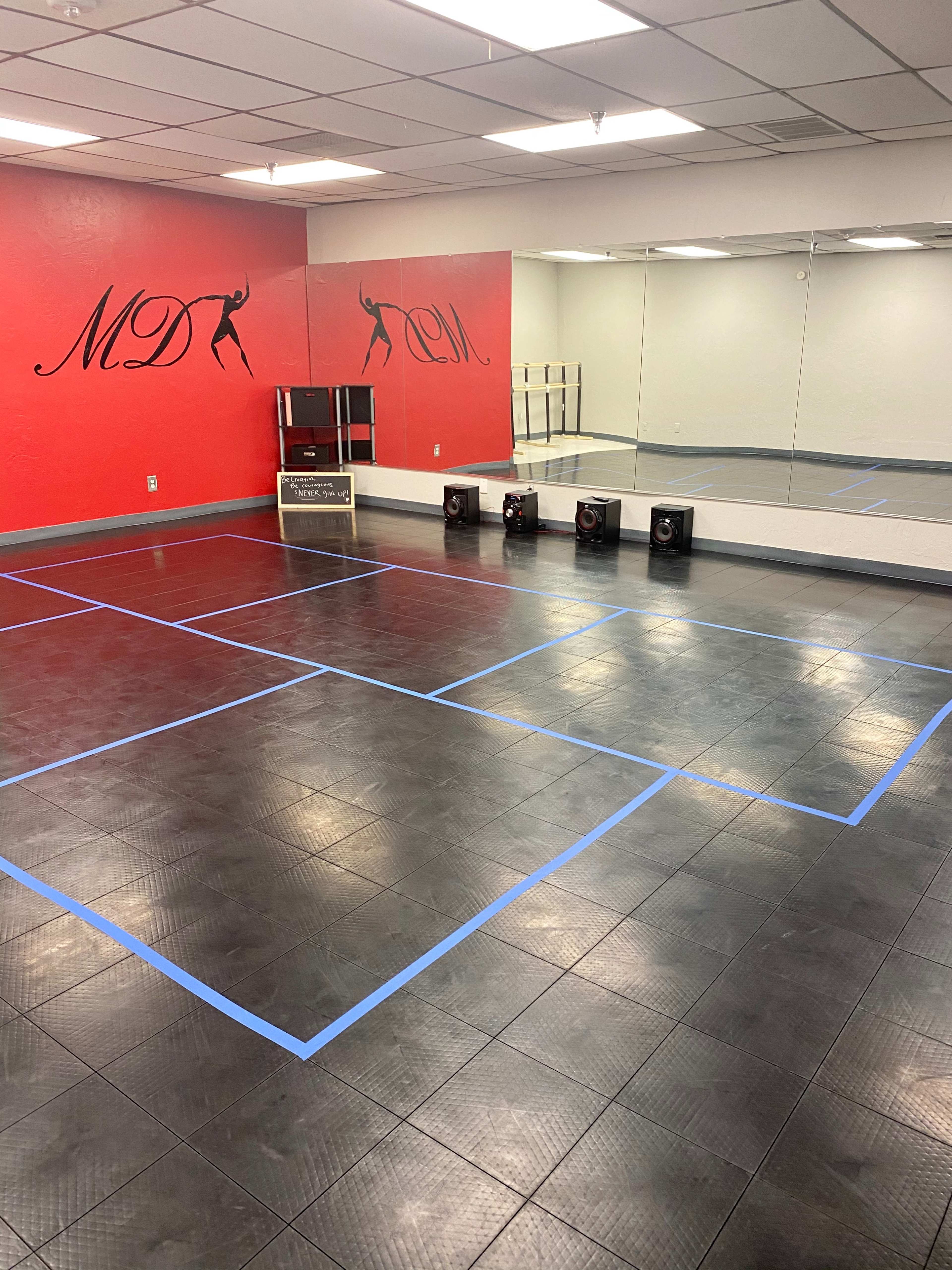 A dance studio with a red and white wall featuring the letters "MD," a mirrored wall, and a black tiled floor marked with blue tape outlining squares.