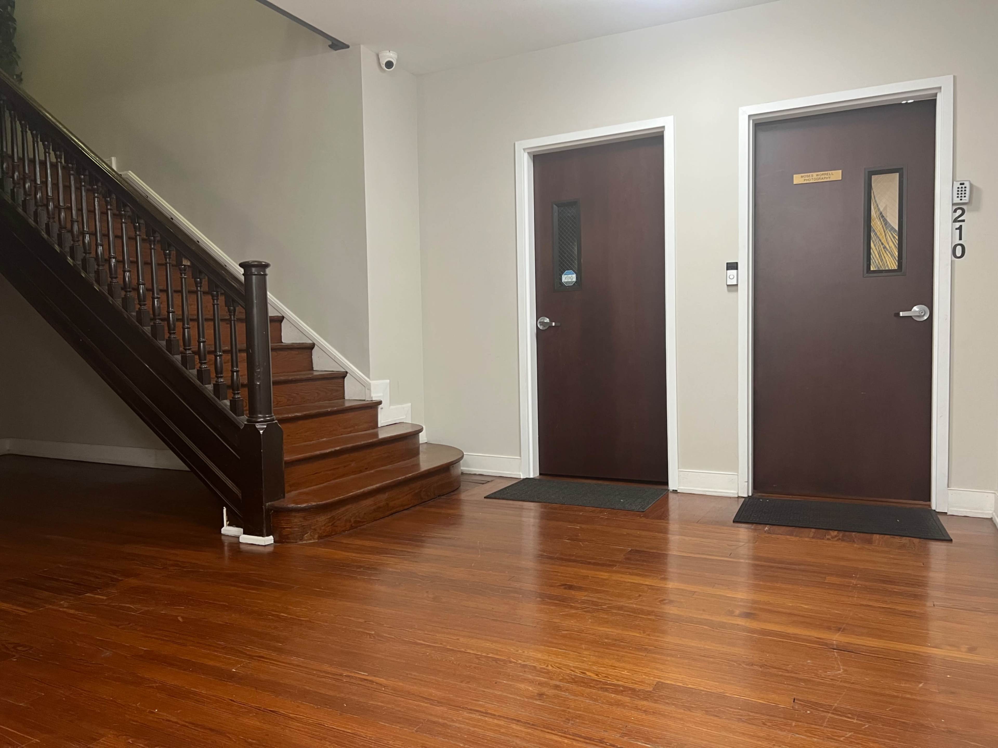 The image shows a hardwood-floored hallway with a staircase on the left and two closed doors on the right, labeled with the numbers 210.