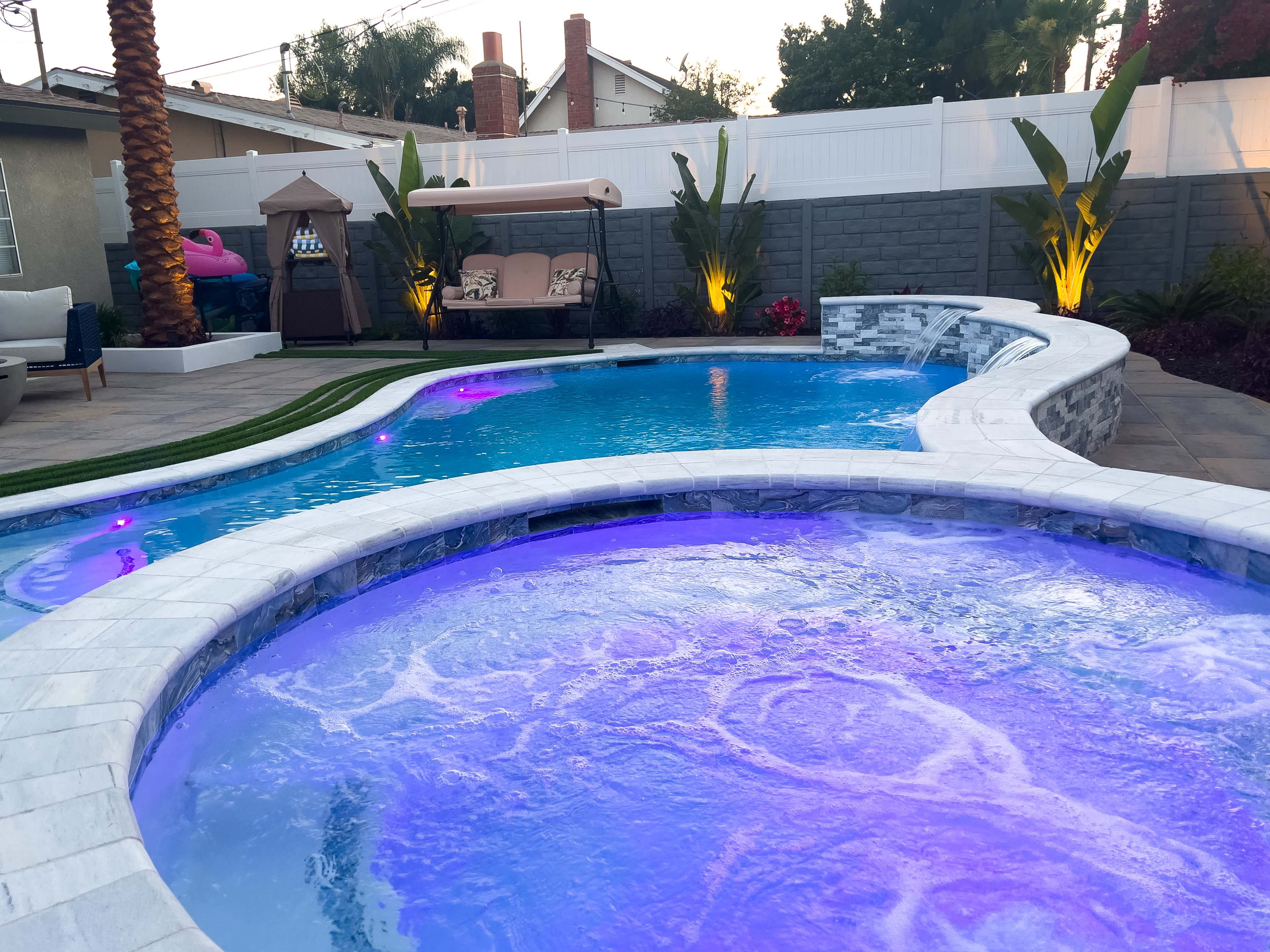A backyard pool area featuring a curved pool with blue water, a spa with purple lighting, and lounge chairs surrounded by tropical plants.