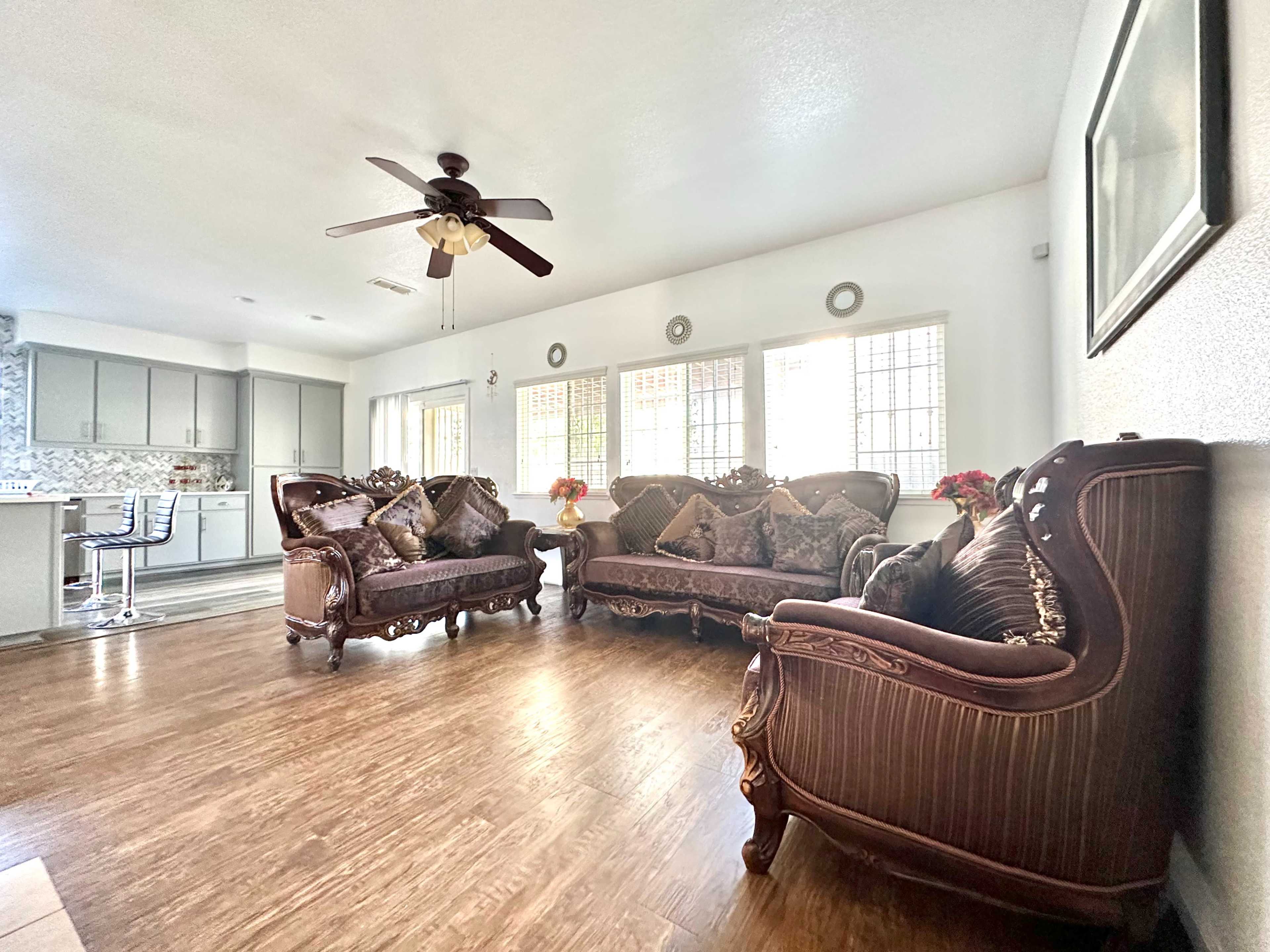 The image shows a living room furnished with ornate brown sofas and a ceiling fan, illuminated by natural light from large windows.