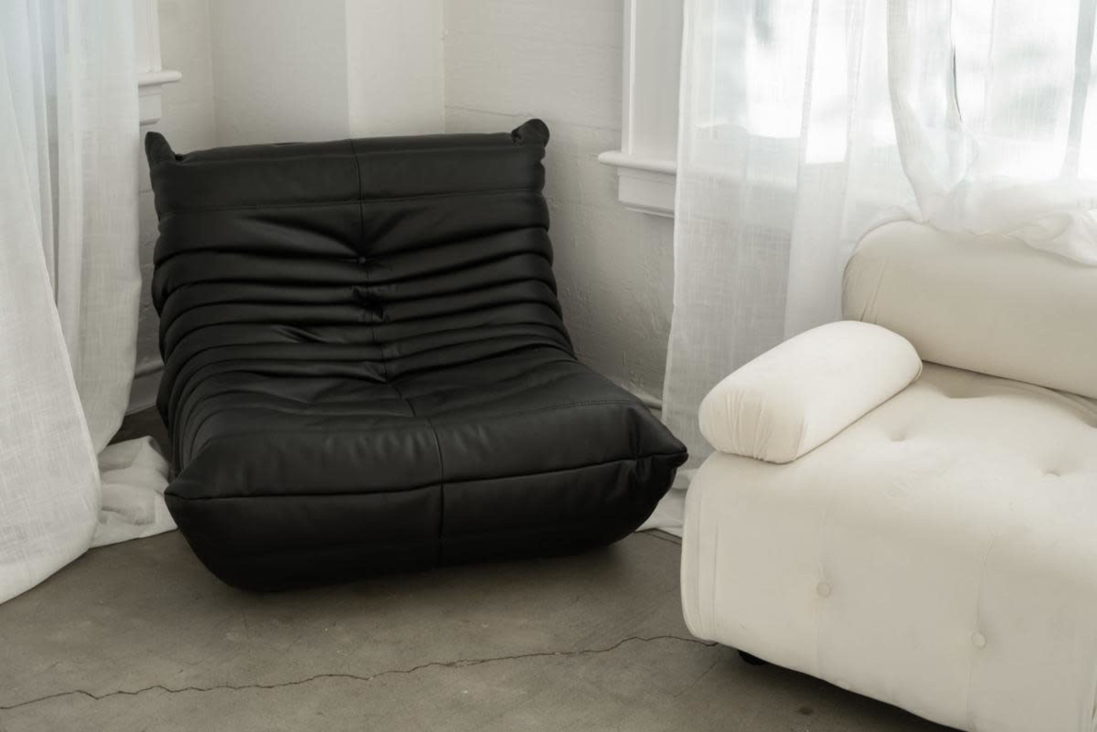 The image shows a black, modern lounge chair with a textured surface positioned alongside a white, tufted armchair in a bright room with sheer curtains.