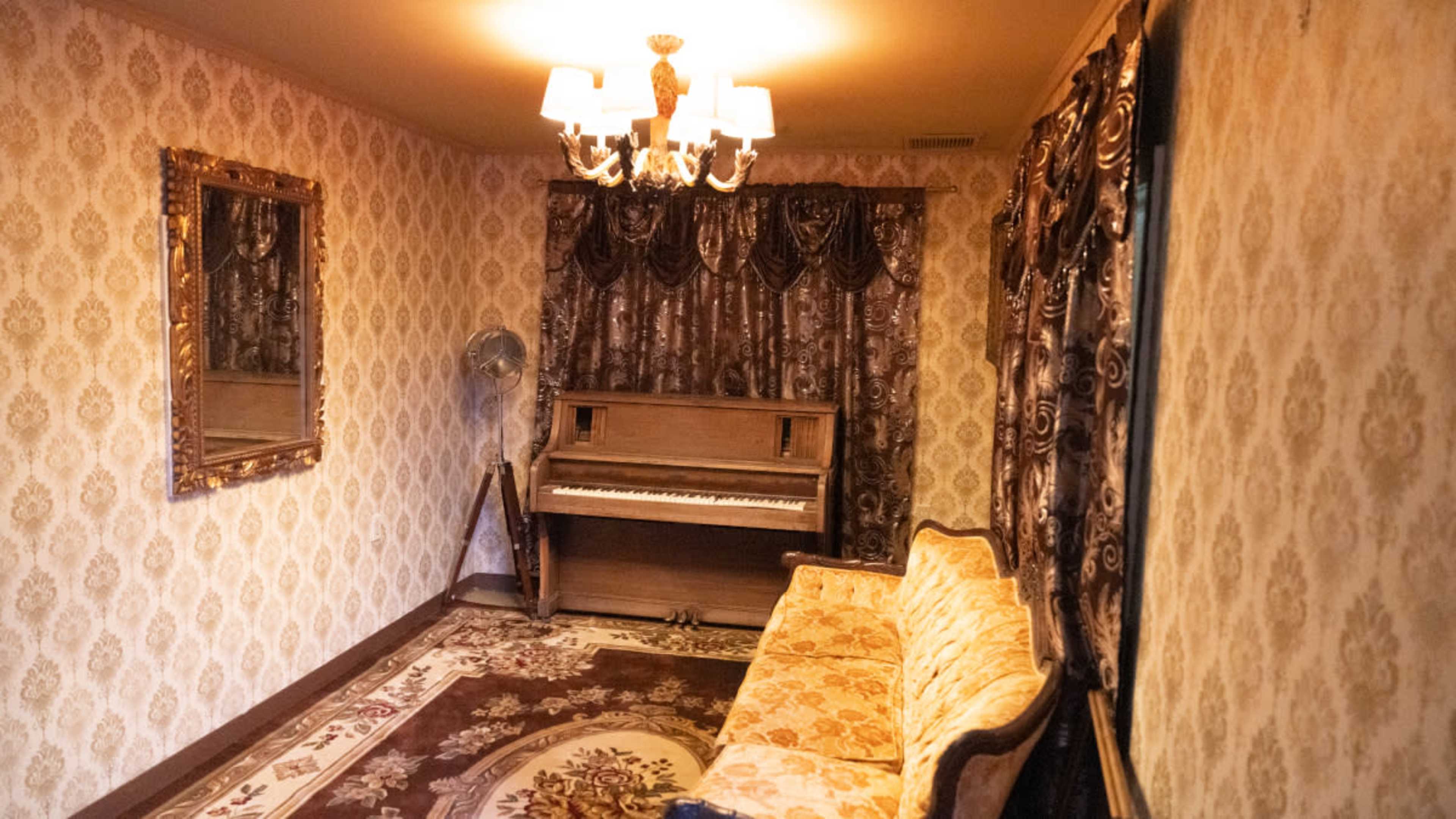 The room features a vintage piano, an ornate mirror, and a decorative sofa, all set against patterned wallpaper and drapes.