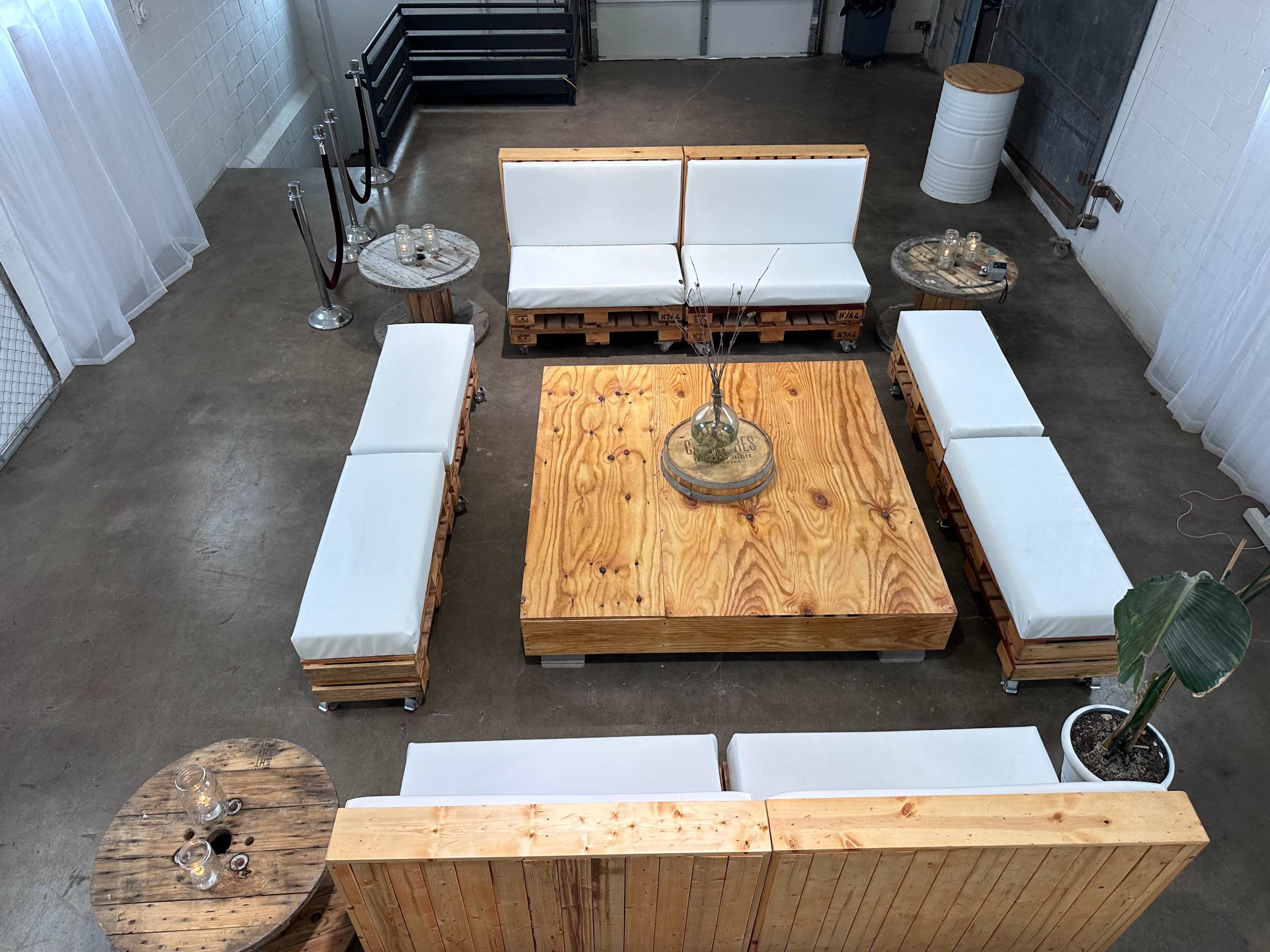 The image shows a spacious indoor area arranged with wooden pallet furniture, including a large square table surrounded by seating and small side tables.