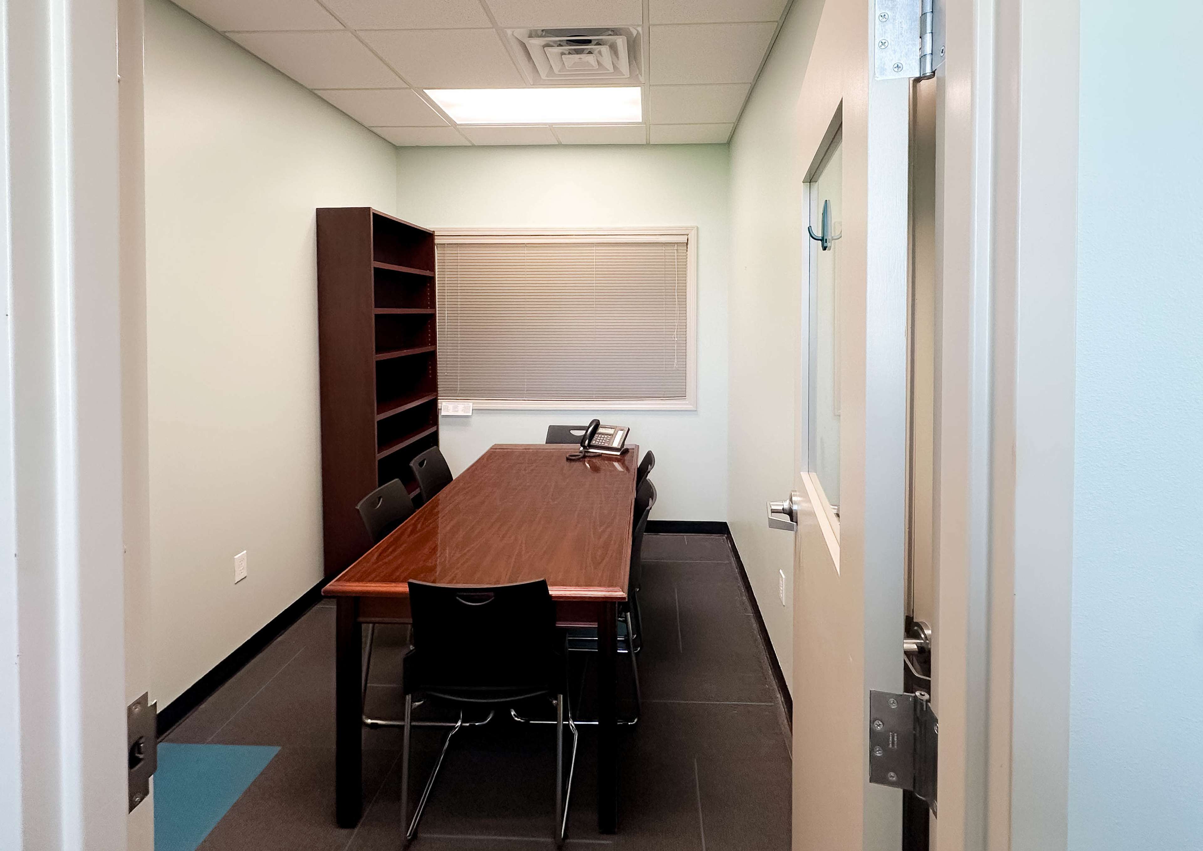 A small conference room features a long wooden table surrounded by black chairs, with a bookshelf and a window on one wall.