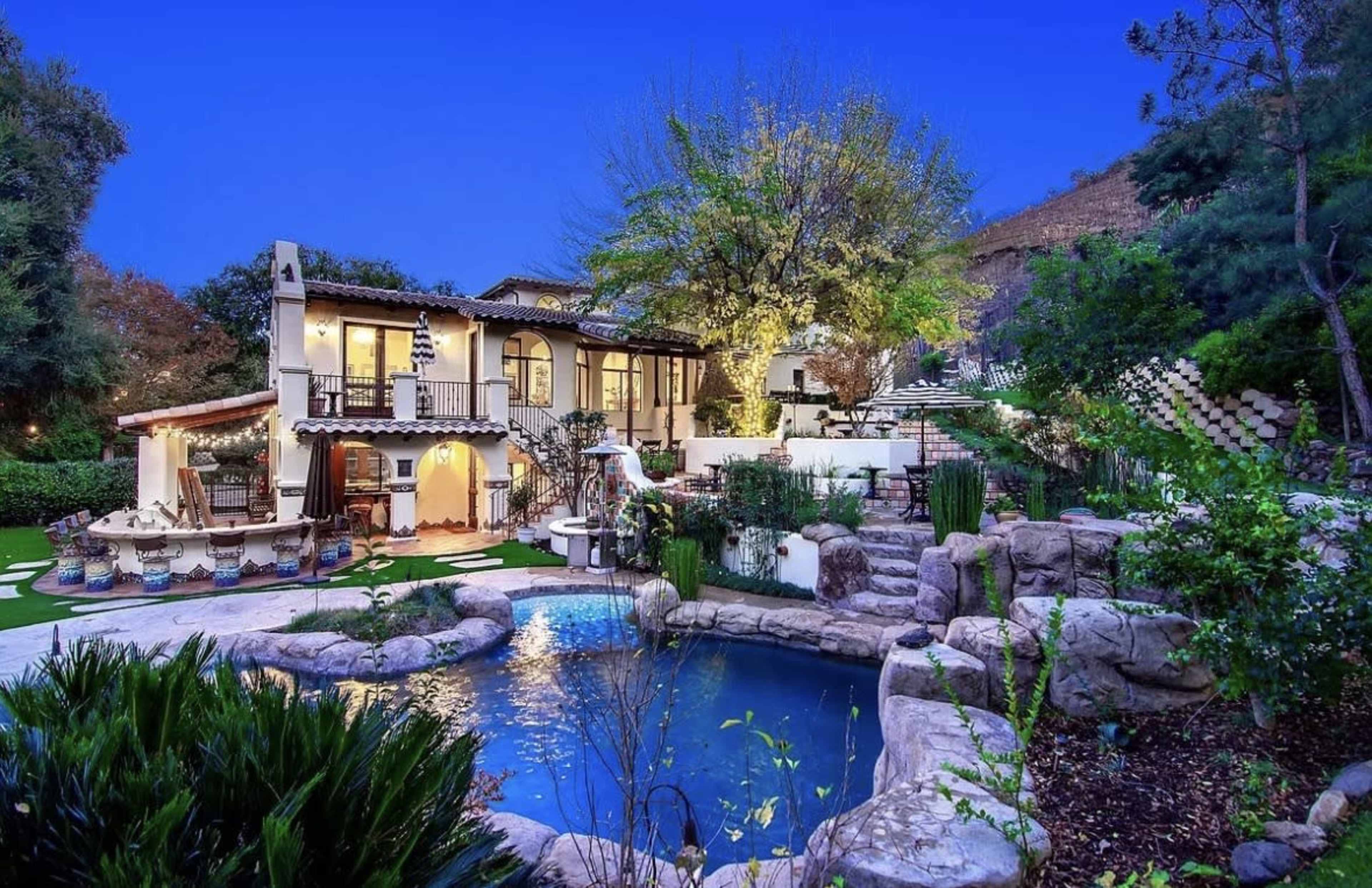 The image shows a beautifully landscaped backyard with a large stone pool surrounded by greenery and a two-story house featuring a balcony and outdoor seating area.