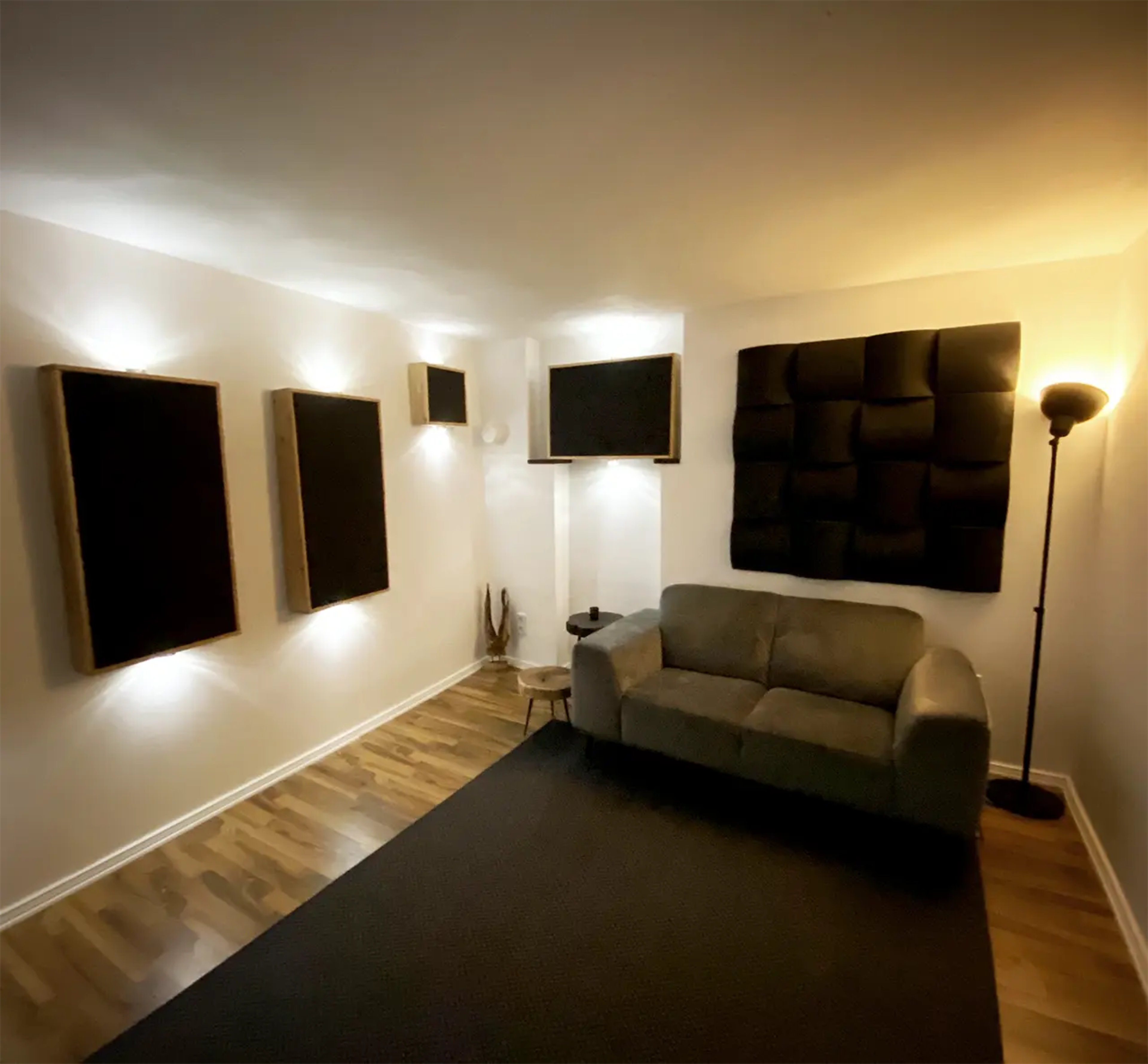 The image depicts a minimalist room featuring a gray sofa, acoustic panels on the walls, and soft lighting.