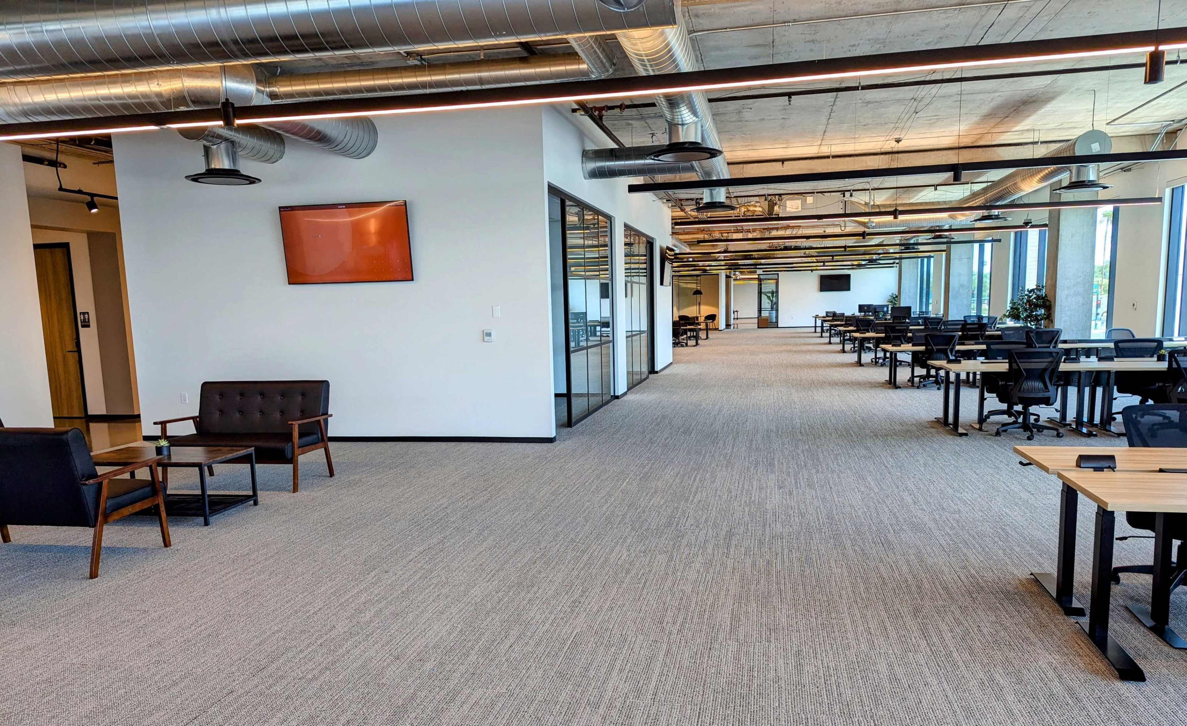 An empty office space featuring long rows of desks, large windows, and a seating area with a couch and a TV on the wall.