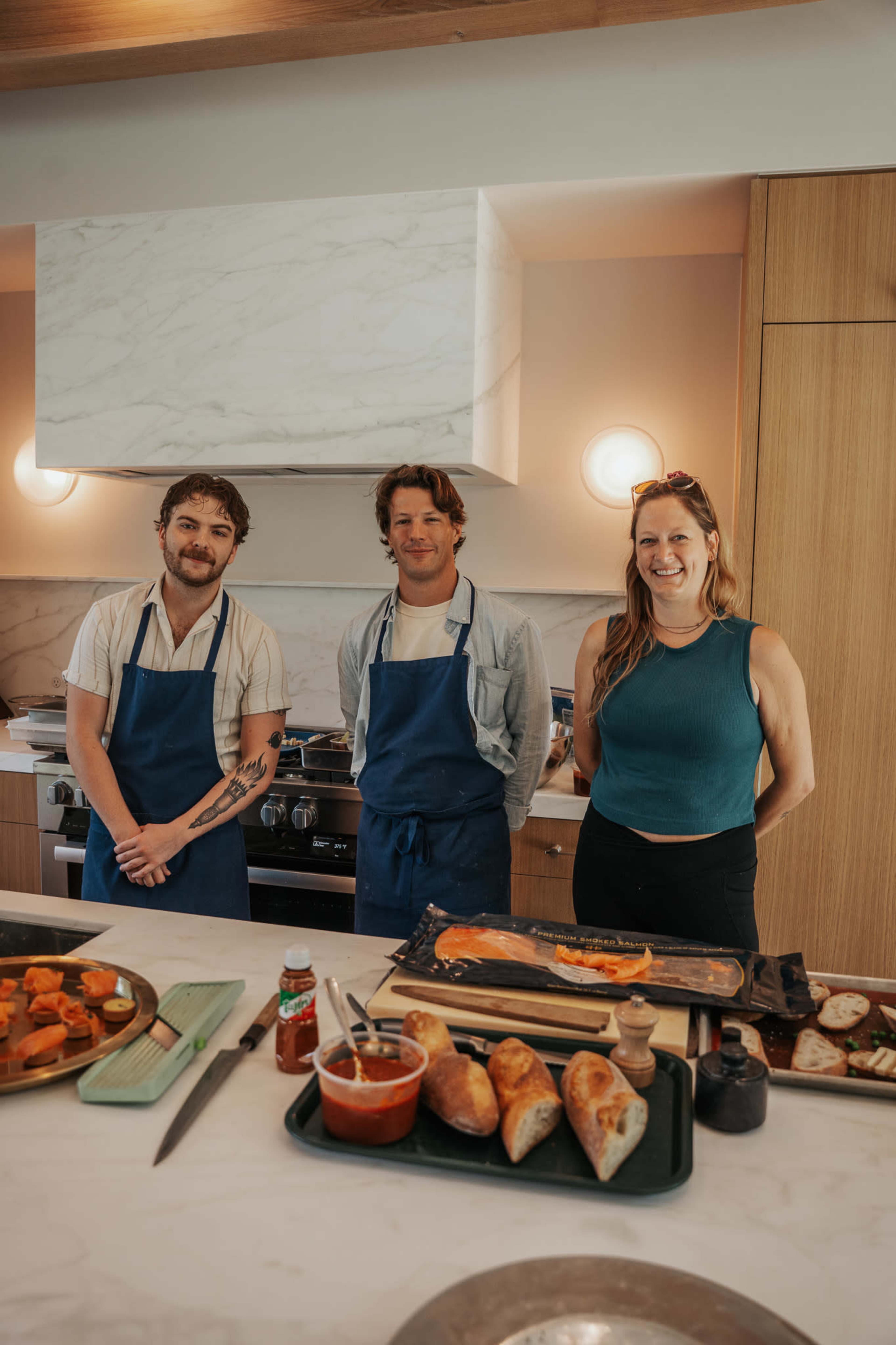 Three individuals, two men and one woman, stand together in a modern kitchen, wearing aprons with various food dishes arranged on the countertop nearby.