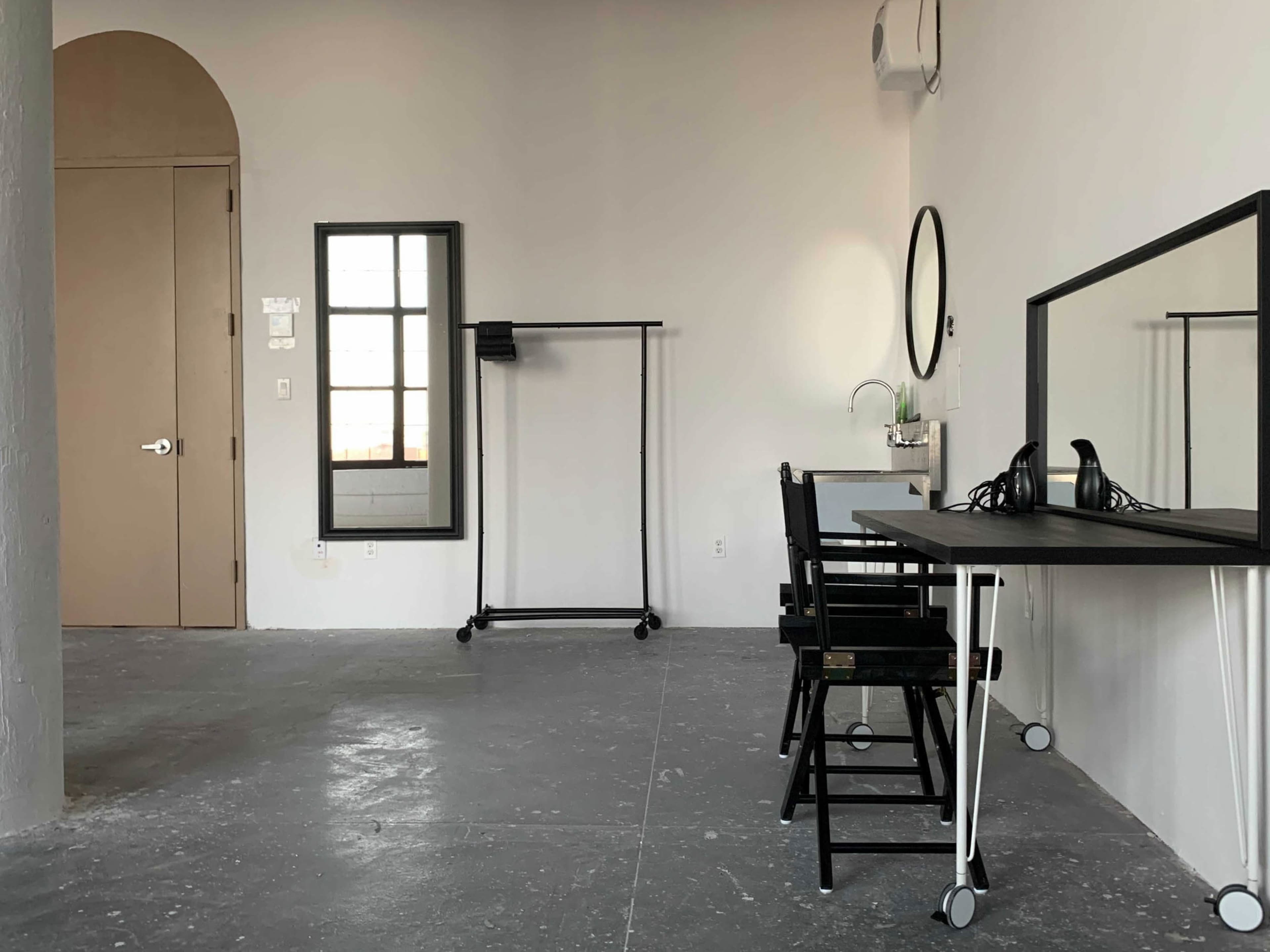 A minimalist room with a desk, a mirror, a clothing rack, and a window in a light-colored interior space.