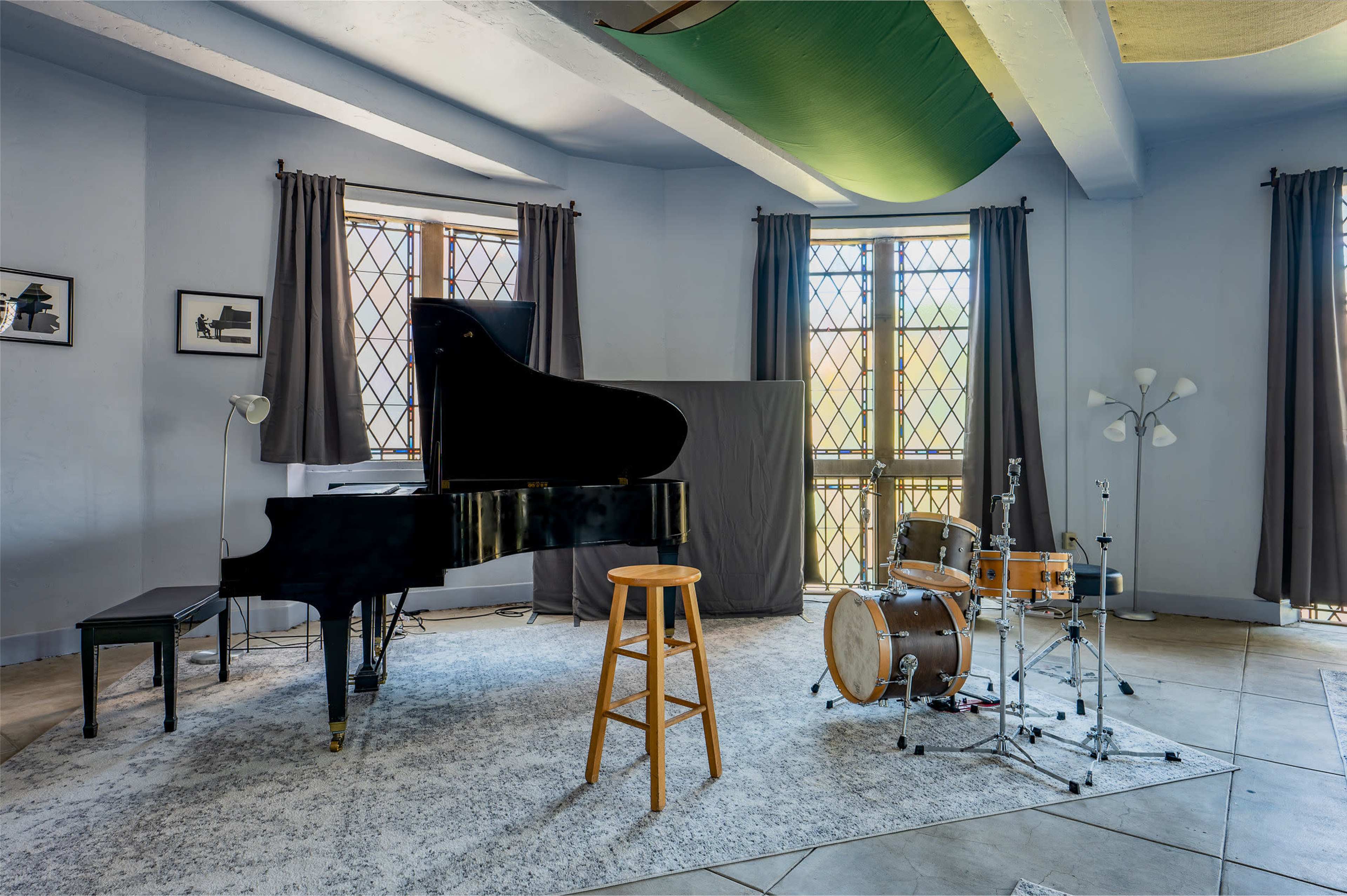 A grand piano and a small drum set are positioned on a rug in a room with large windows and gray curtains.