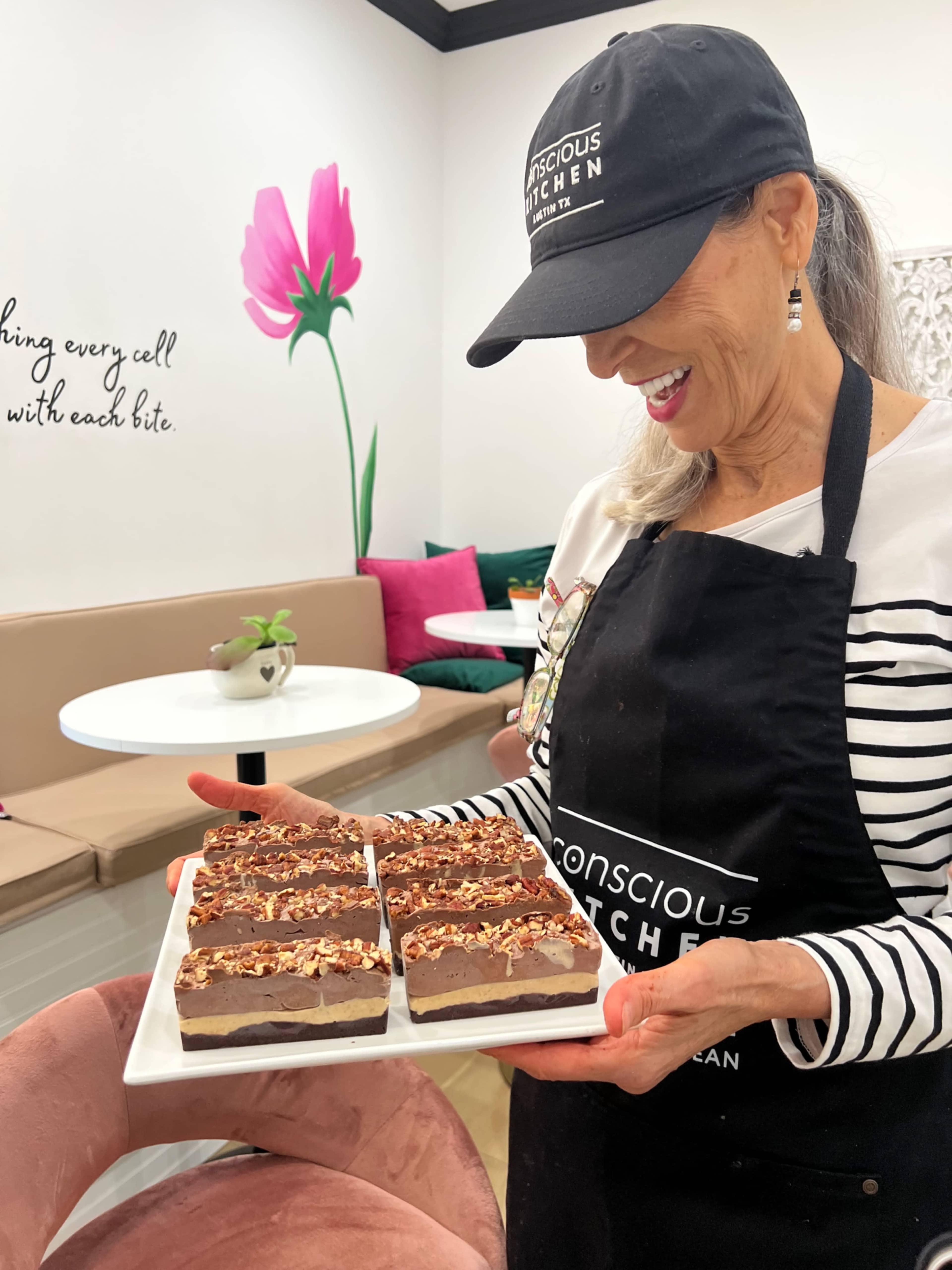 A woman in an apron and cap holds a platter of layered chocolate desserts while smiling in a brightly decorated cafe.