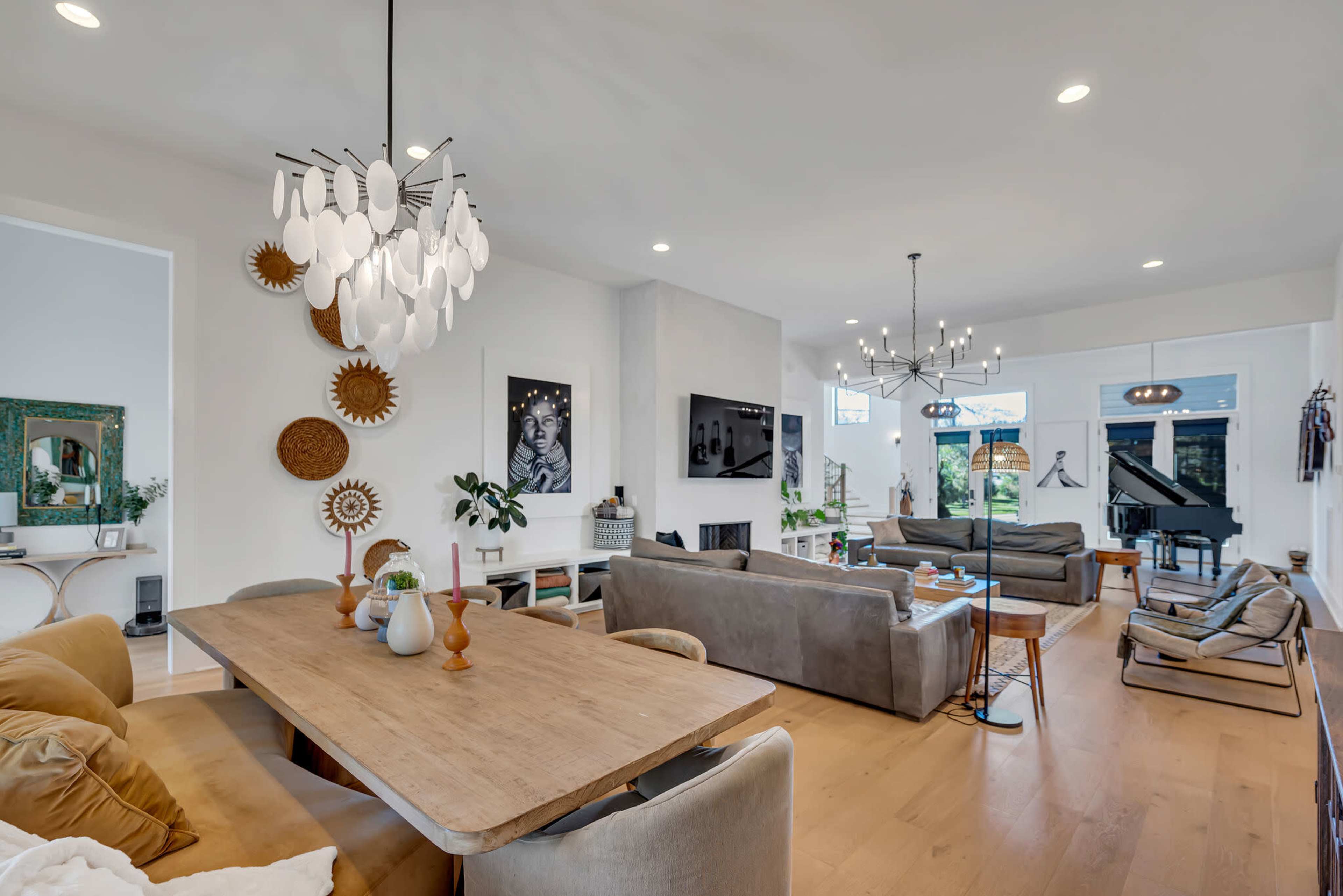 A modern living room features a dining area with a wooden table, comfortable sofas, decorative wall art, and large windows allowing natural light.