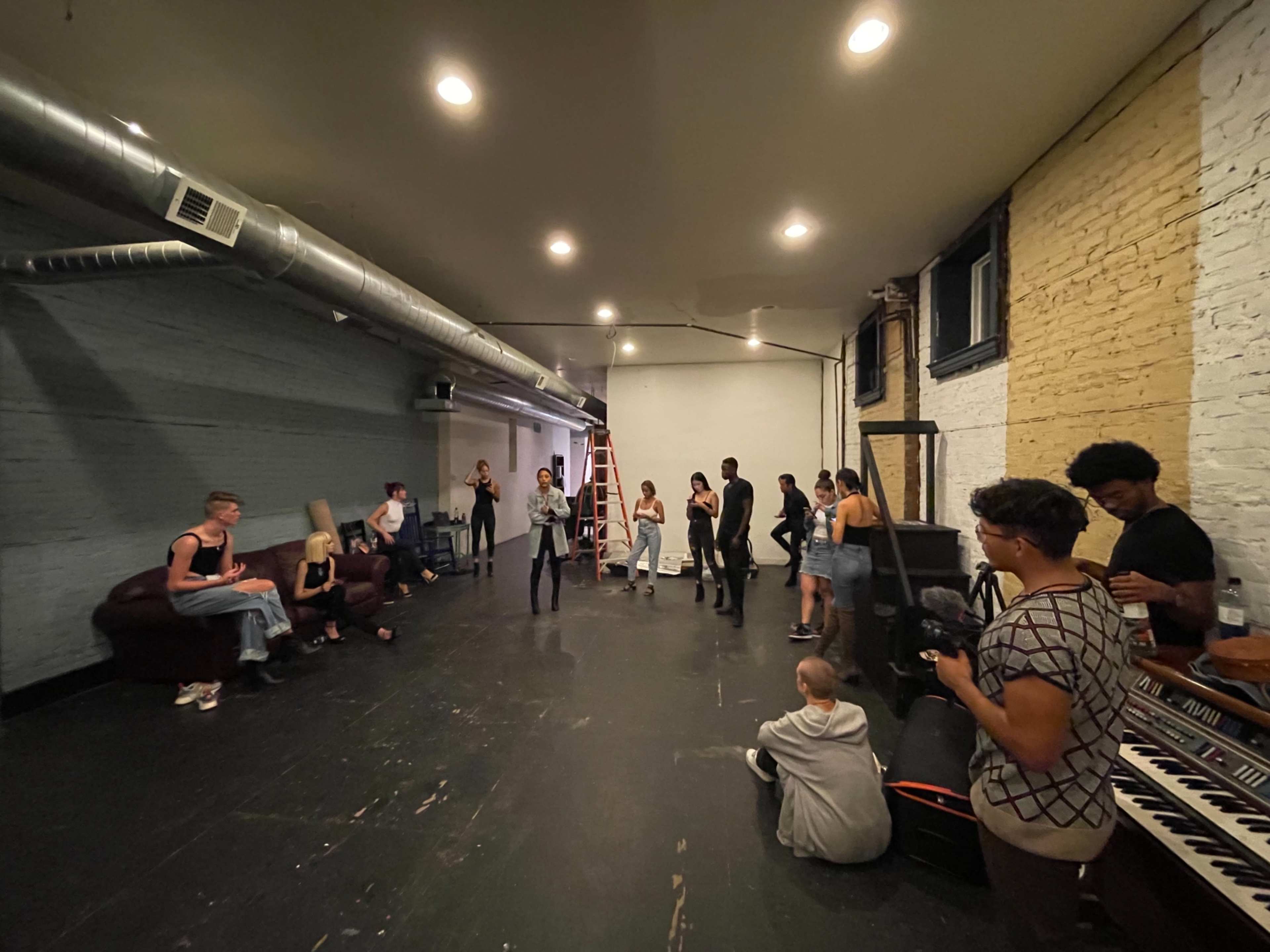 A group of people is gathered in a spacious, industrial-style room with exposed brick walls and hardwood floors, some standing while others are seated.