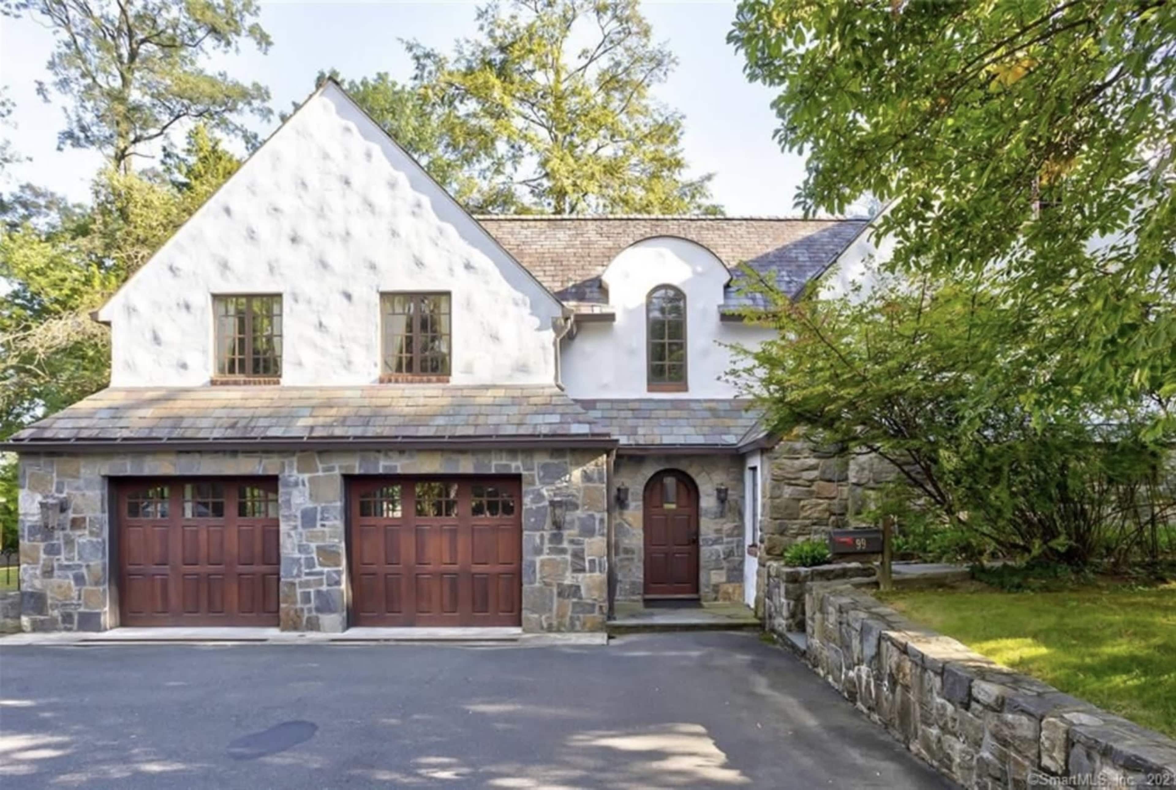 A two-story house with a stone facade and wooden garage doors is set against a backdrop of trees.