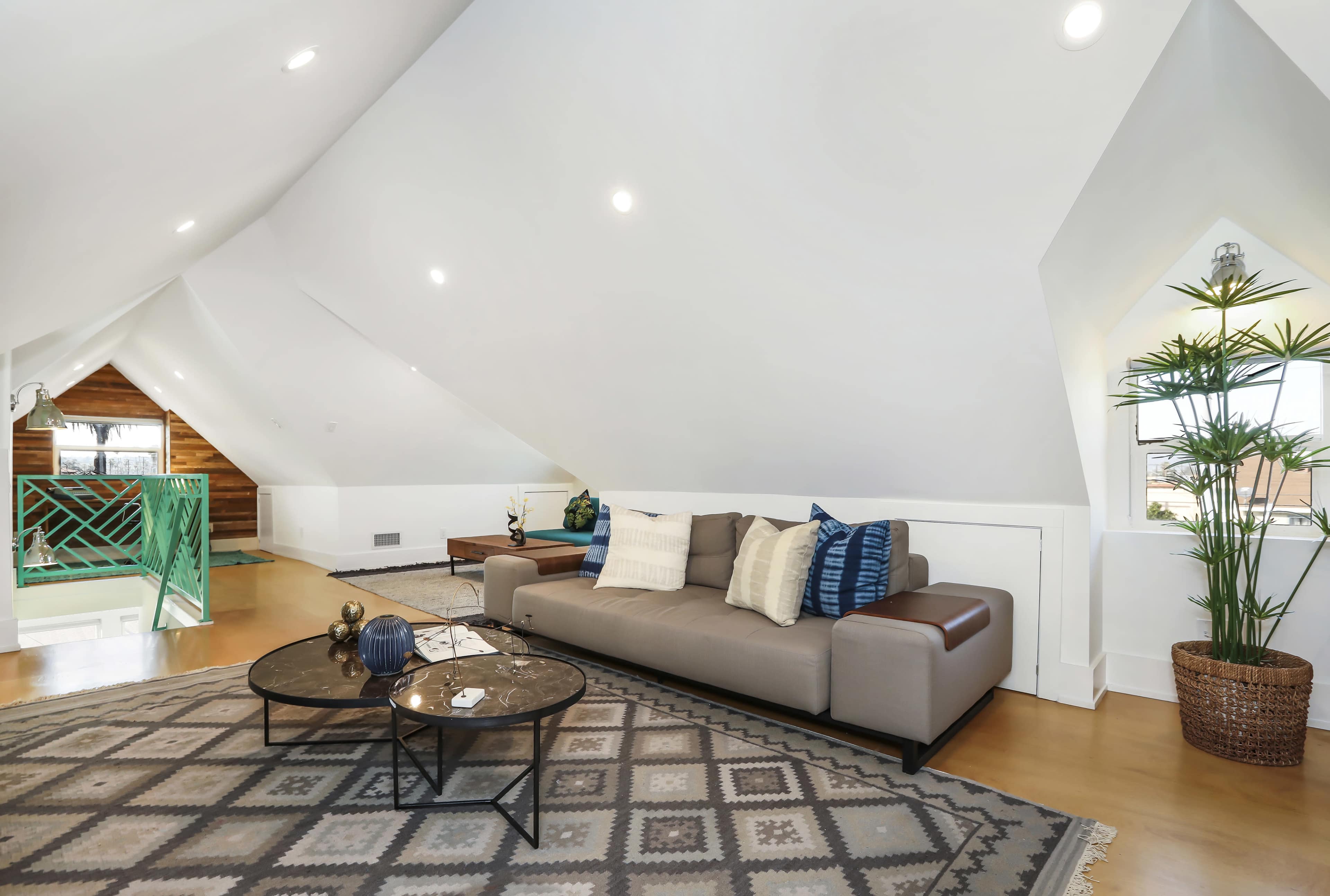 The image shows a modern living room with a sloped ceiling, featuring a light-colored sofa, round coffee tables, and a potted plant beside a window.